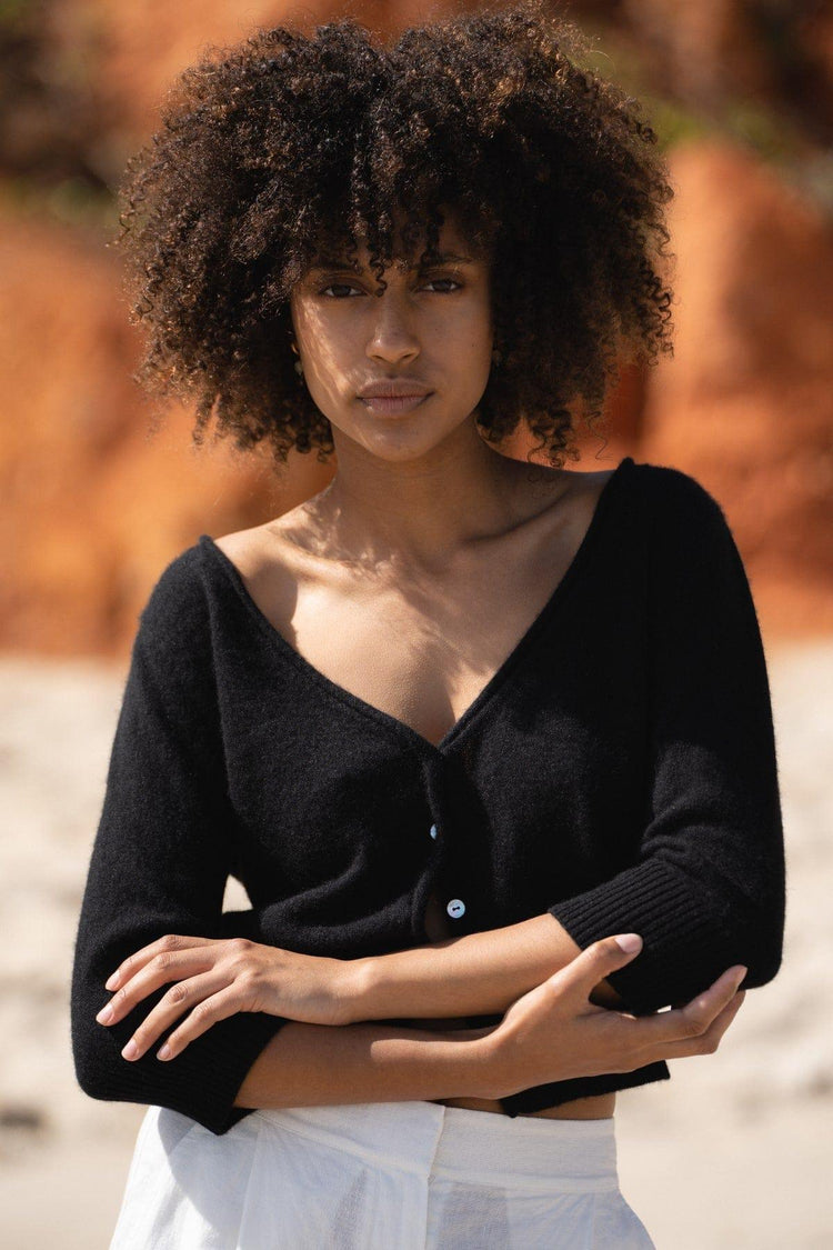 Jade Cashmere Cardigan in Black - front view, arms crossed, worn with wide-leg trousers, standing on red rock beach - Lilly Pilly Collection
