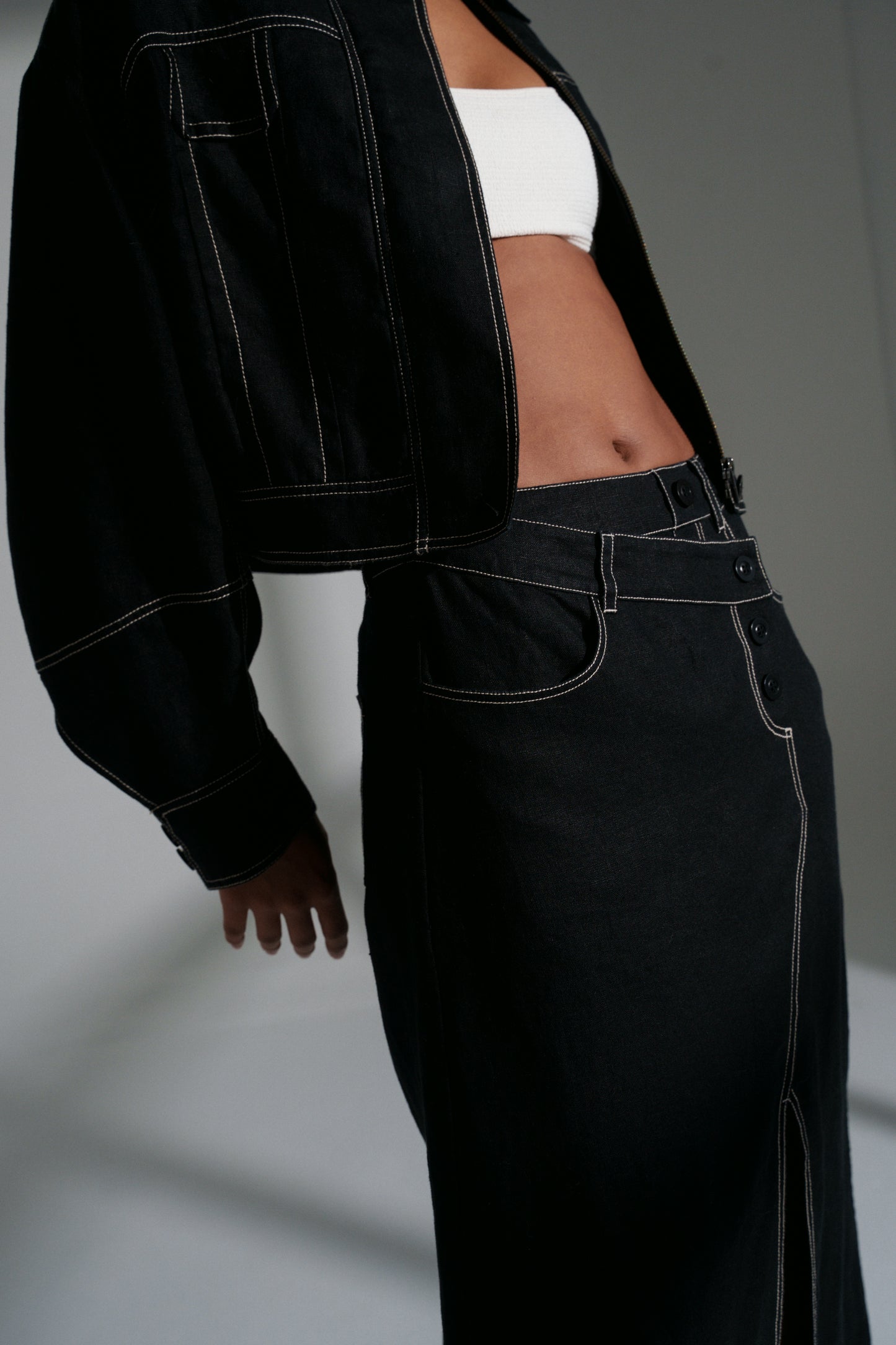 Side view of LILLY PILLY woman wearing a black Wes linen jacket and Jax linen skirt standing against a neutral background.