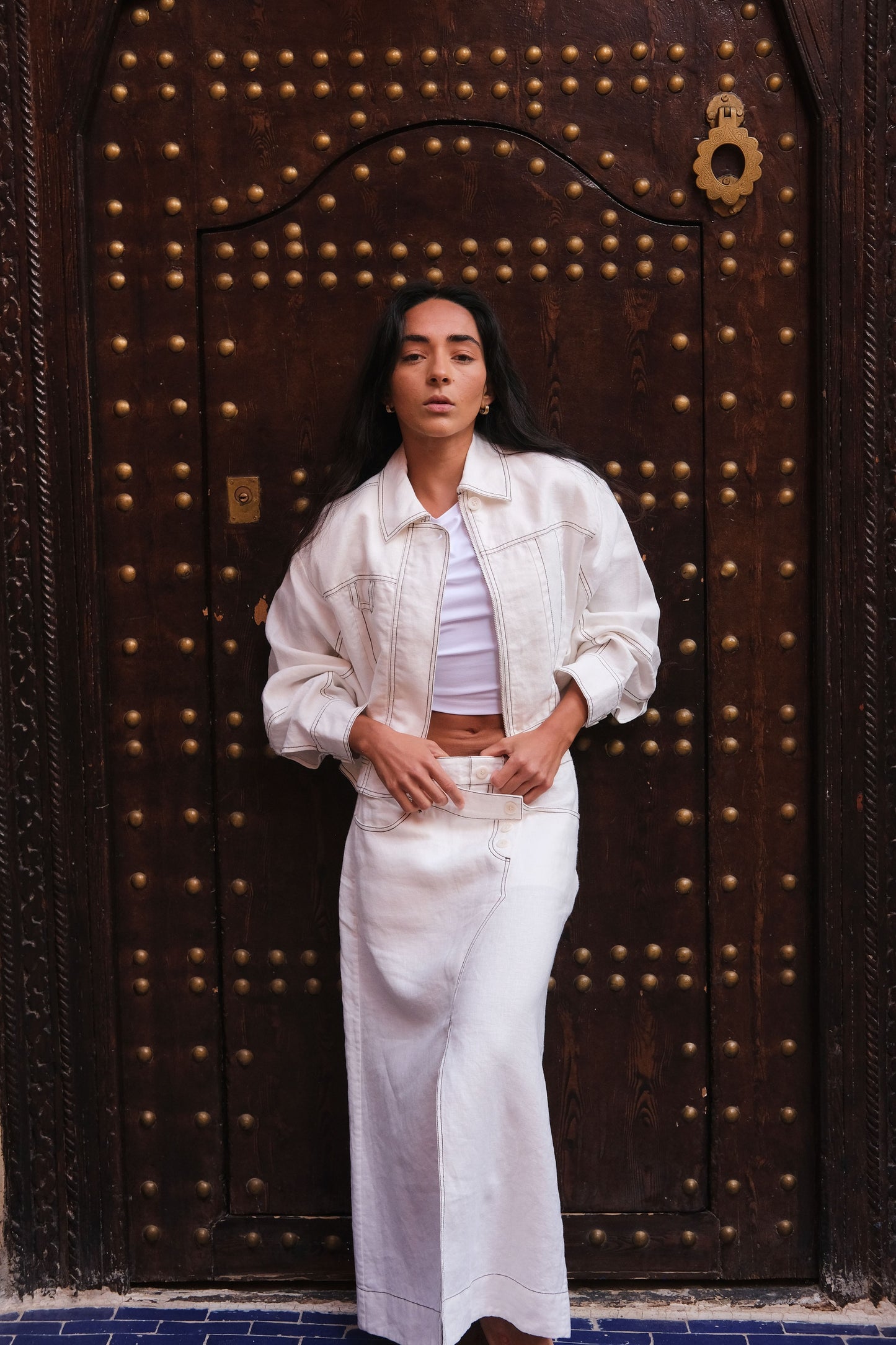 LILLY PILLY woman in a ivory Jax Skirt and Wes Jacket standing in front of a large wooden door with metallic studs.