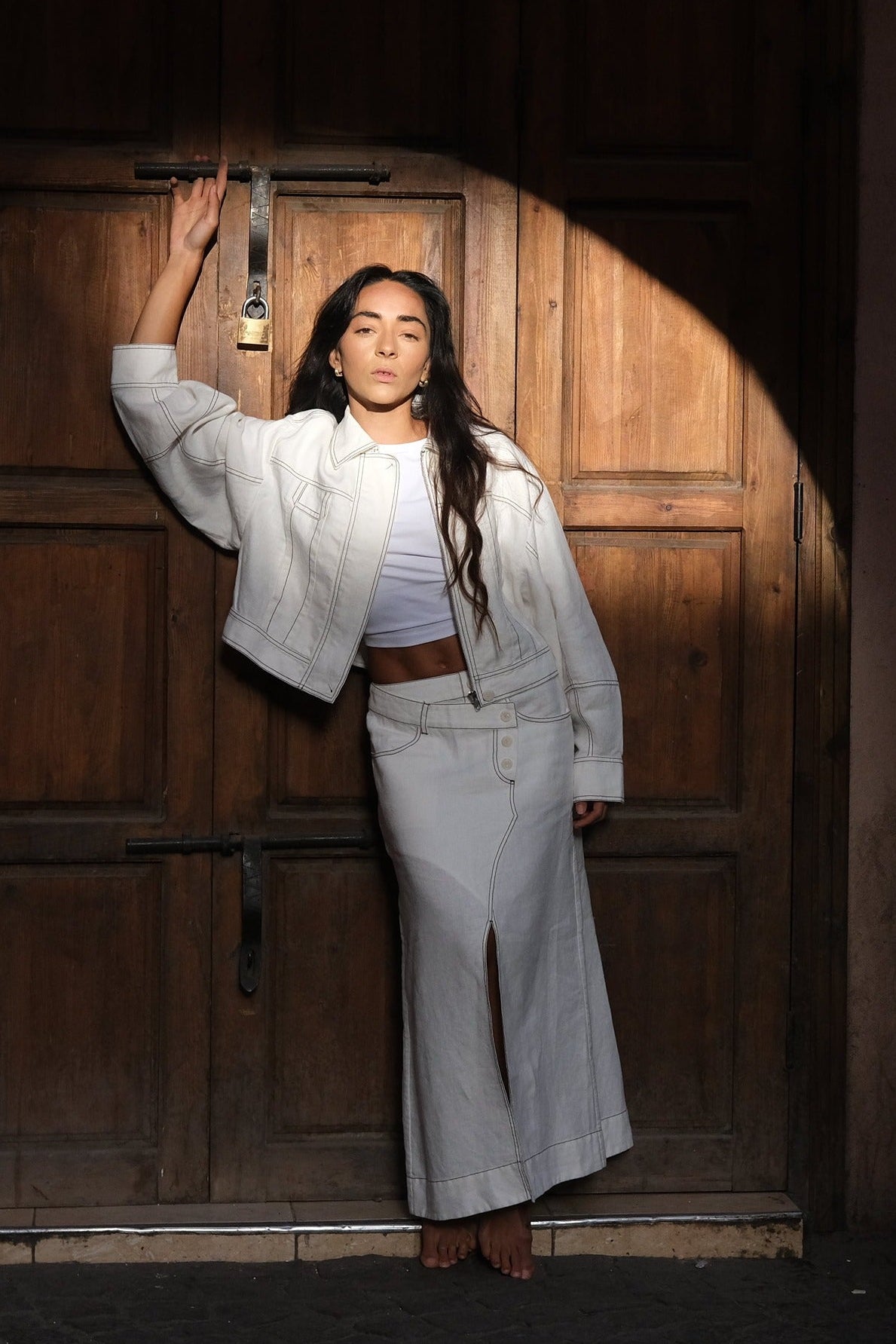 LILLY PILLY woman in a ivory Jax Skirt and Wes Jacket standing in front of a wooden door with a warm light.