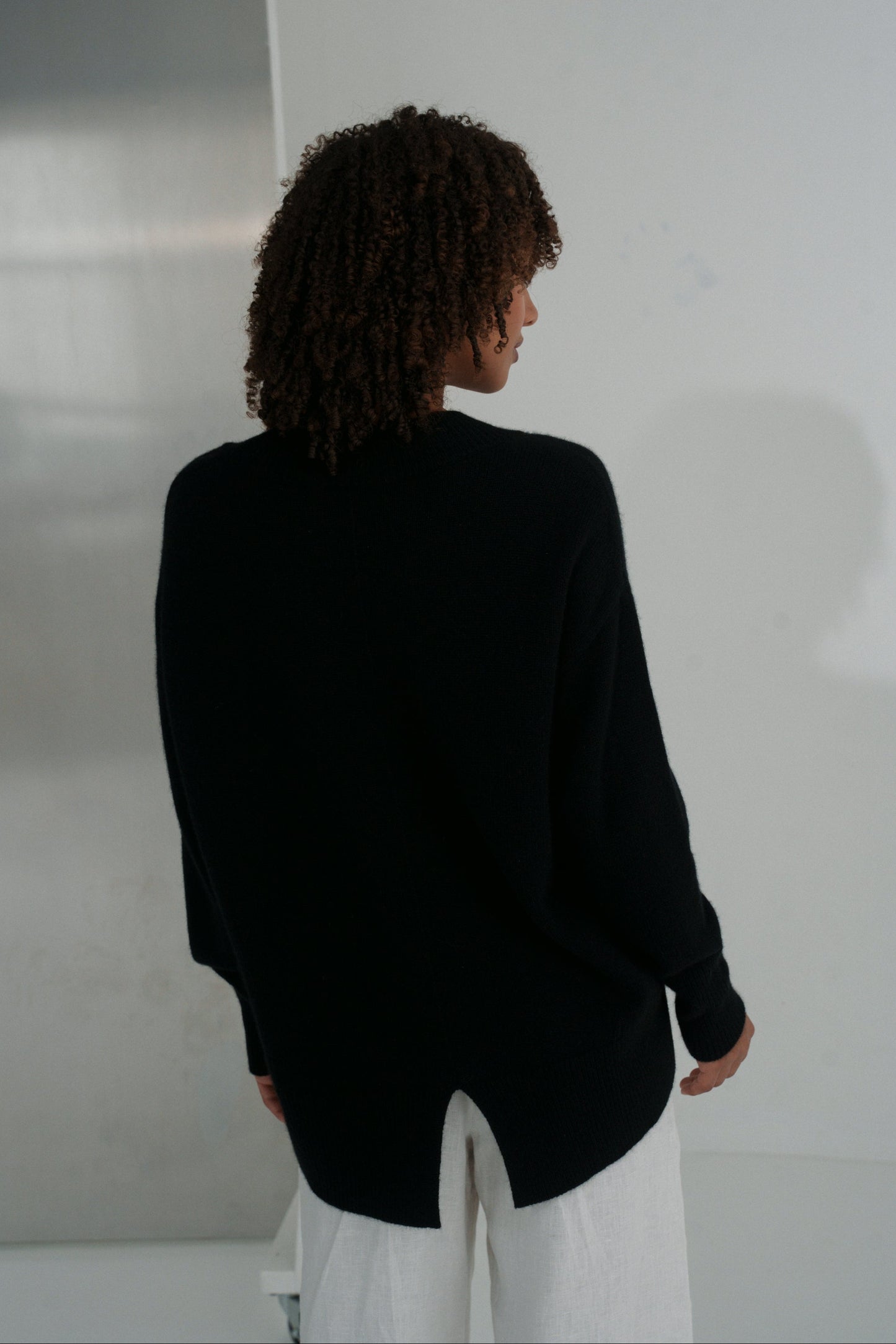 Back view of LILLY PILLY woman wearing an black June cashmere knit and ivory linen pants against a plain background