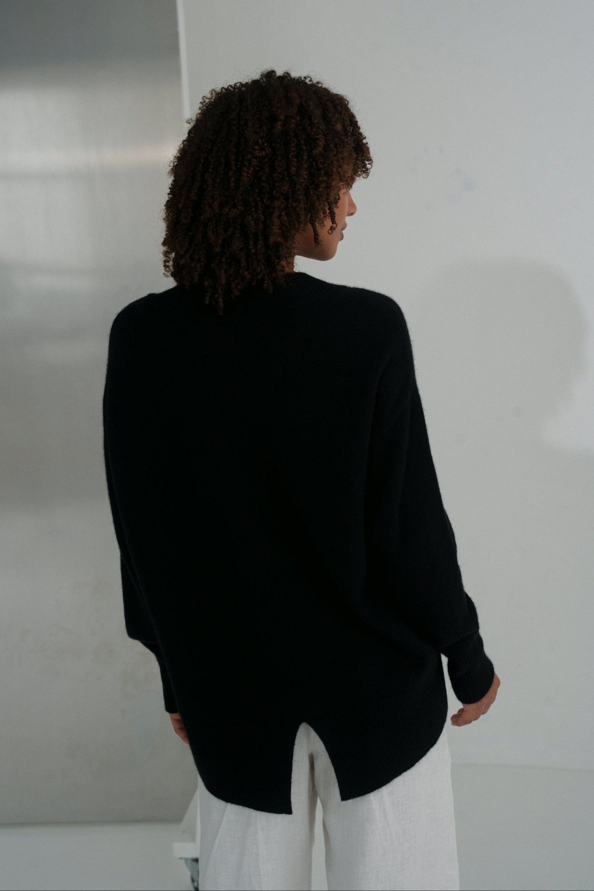 Back view of LILLY PILLY woman wearing an black June cashmere knit and ivory linen pants against a plain background