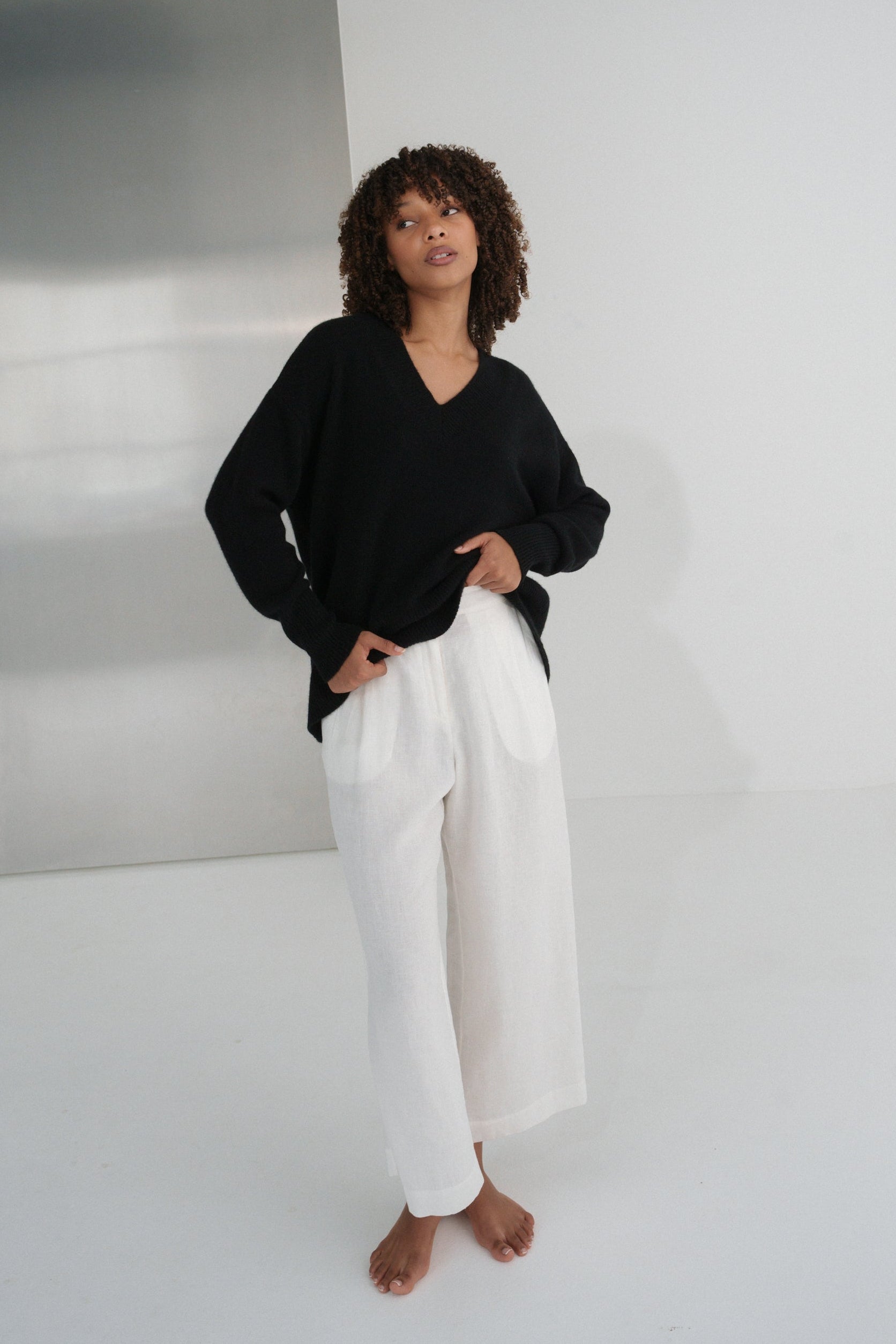 LILLY PILLY woman wearing an black June cashmere knit and ivory linen pants standing against a plain wall.