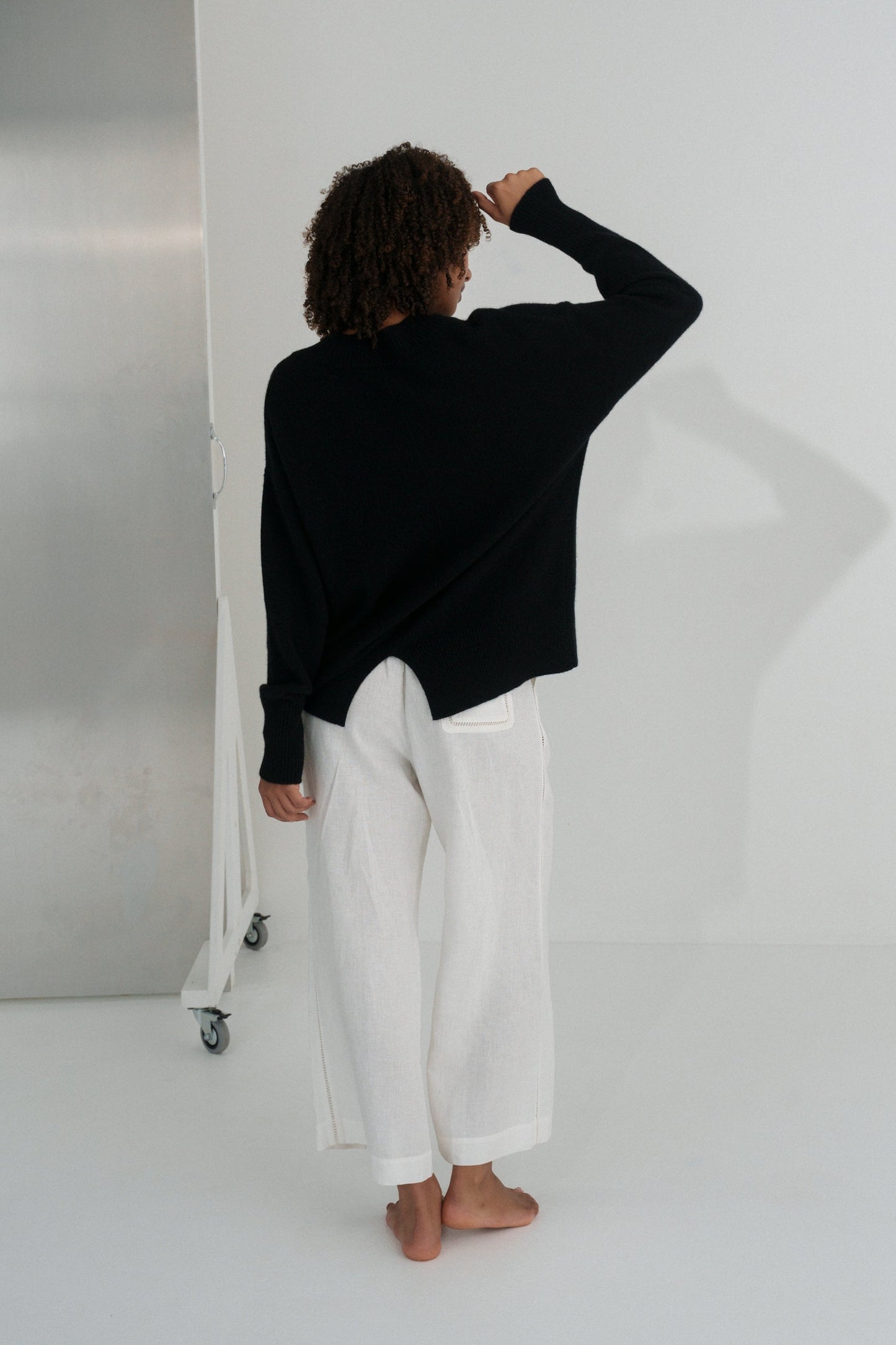 Back view of LILLY PILLY woman wearing an black June cashmere knit and ivory linen pants standing in a minimalistic room.