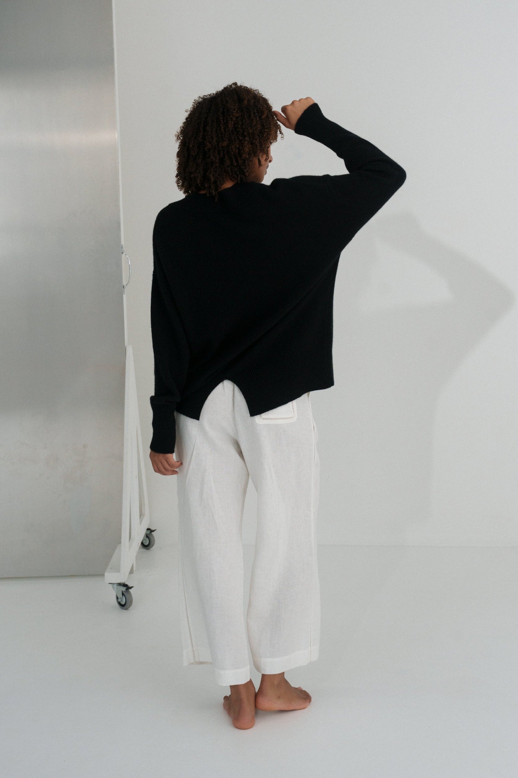 Back view of LILLY PILLY woman wearing an black June cashmere knit and ivory linen pants standing in a minimalistic room.