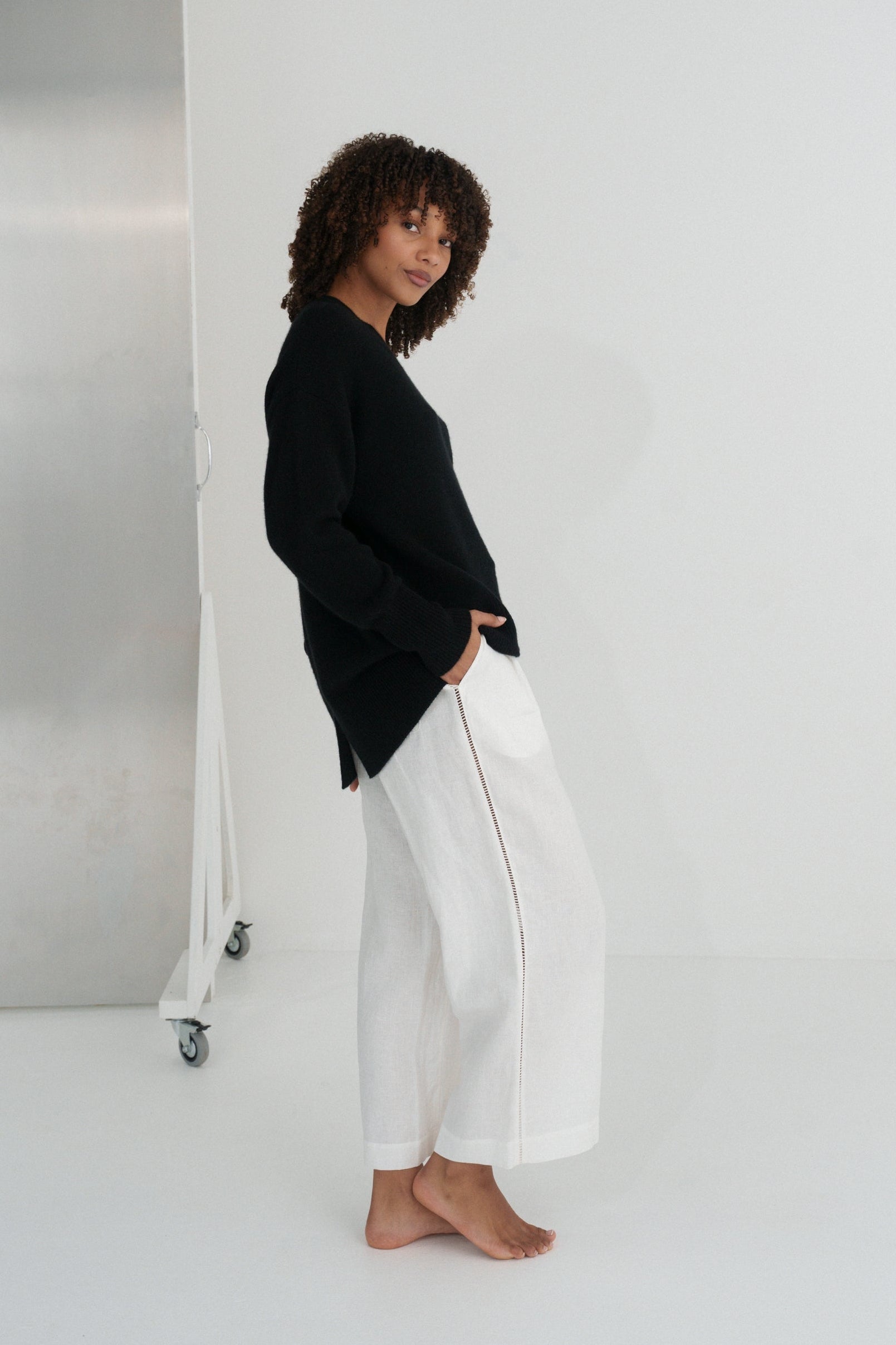 Side view of LILLY PILLY woman wearing an black June cashmere knit and ivory linen pants standing against a white wall.