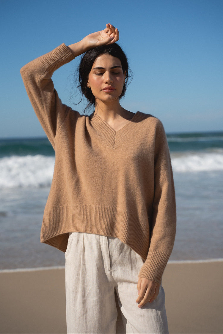 June Cashmere Knit in Camel Brown - front view, arm raised shading eyes, worn with wide-leg trousers, standing on beach with ocean backdrop - Lilly Pilly Collection