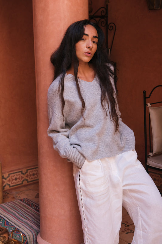 LILLY PILLY woman wearing a grey marle June cashmere knit sweater and ivory linen pants standing against a pillar and orange wall.