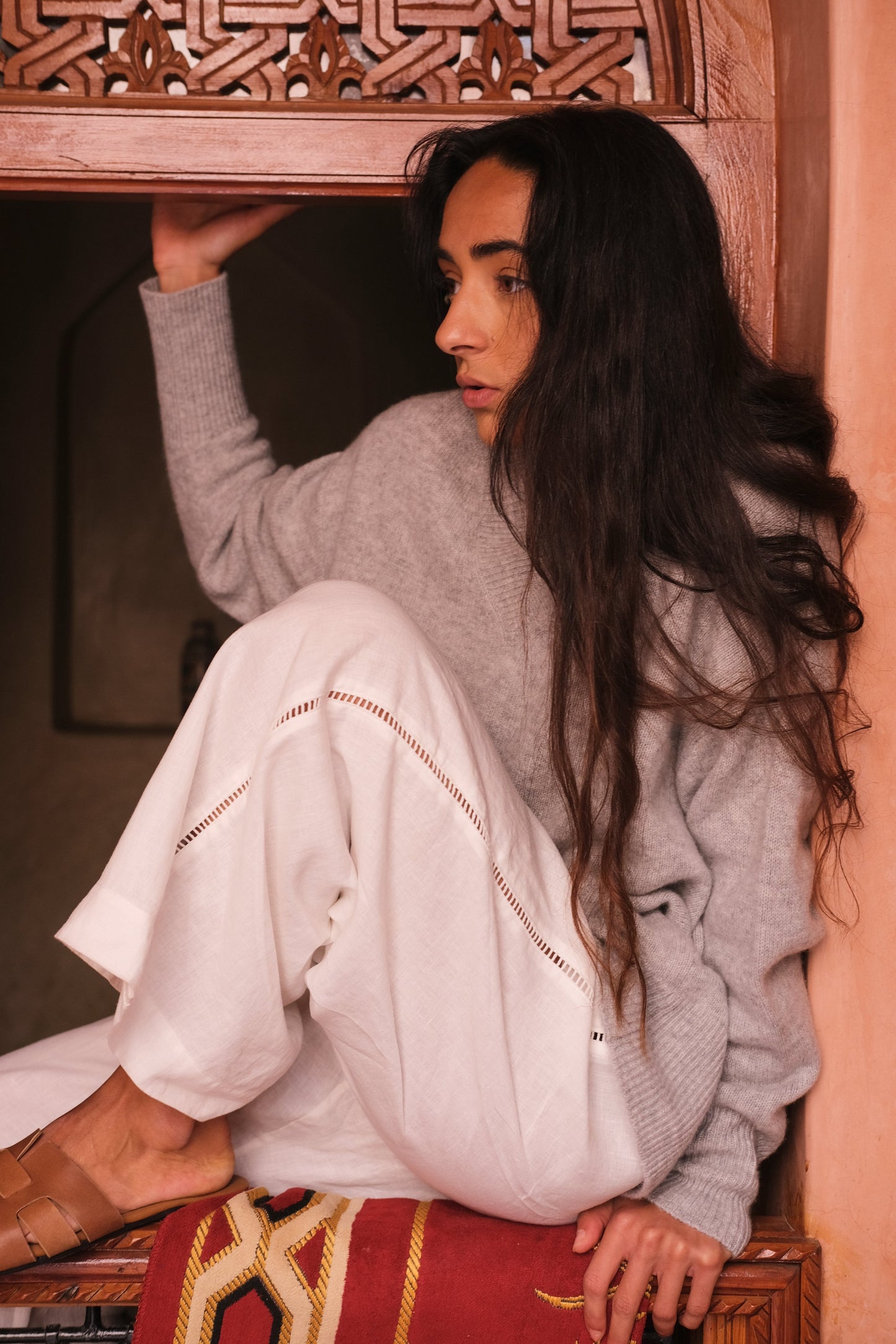 LILLY PILLY woman wearing a grey marle June cashmere knit sweater and ivory linen pants sitting on a wooden bench with intricate carvings