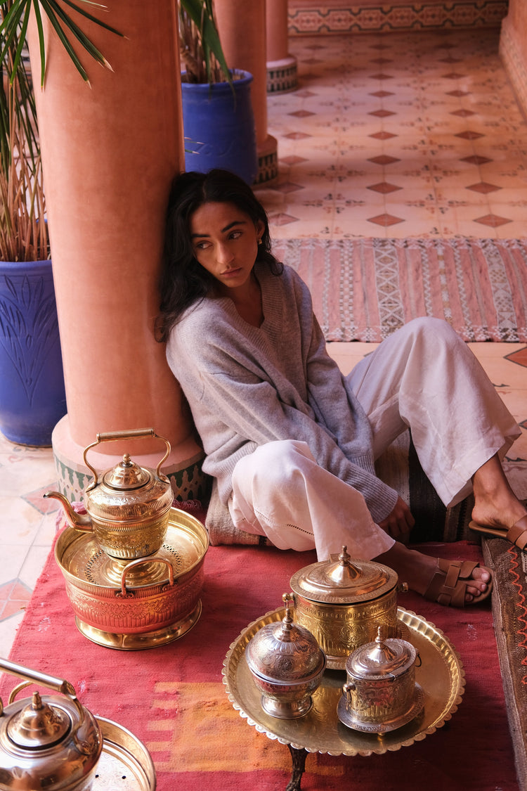 June Cashmere Knit in Grey Marle - front view, styled with ivory linen pants, seated on a patterned rug with decorative brass containers nearby - Lilly Pilly Collection