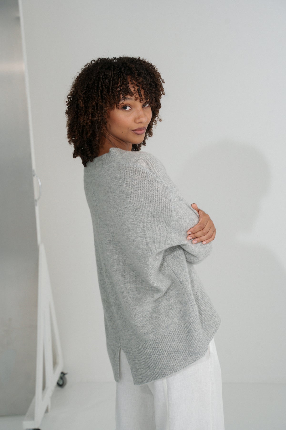 Side view of LILLY PILLY woman wearing an grey marle June cashmere knit sweater and ivory linen pants against a white background