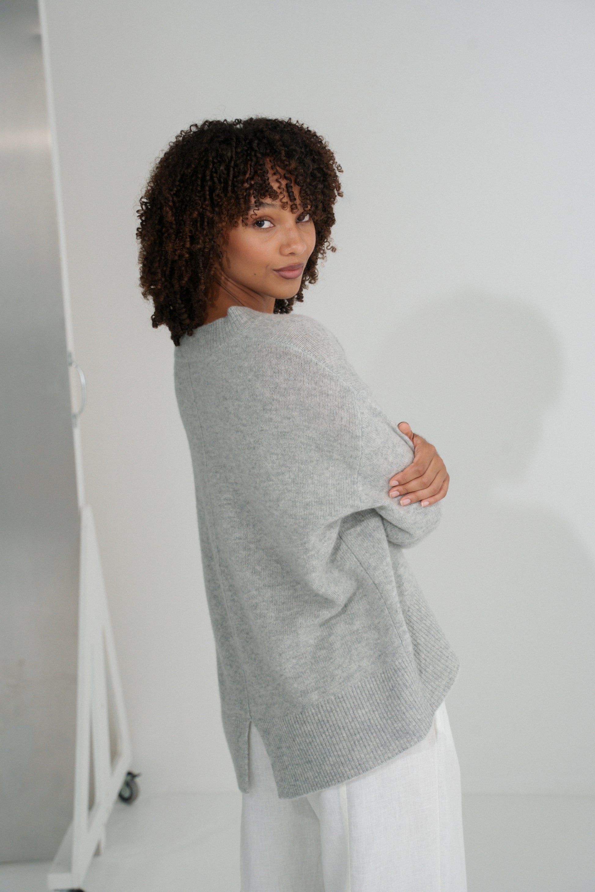 Side view of LILLY PILLY woman wearing an grey marle June cashmere knit sweater and ivory linen pants against a white background
