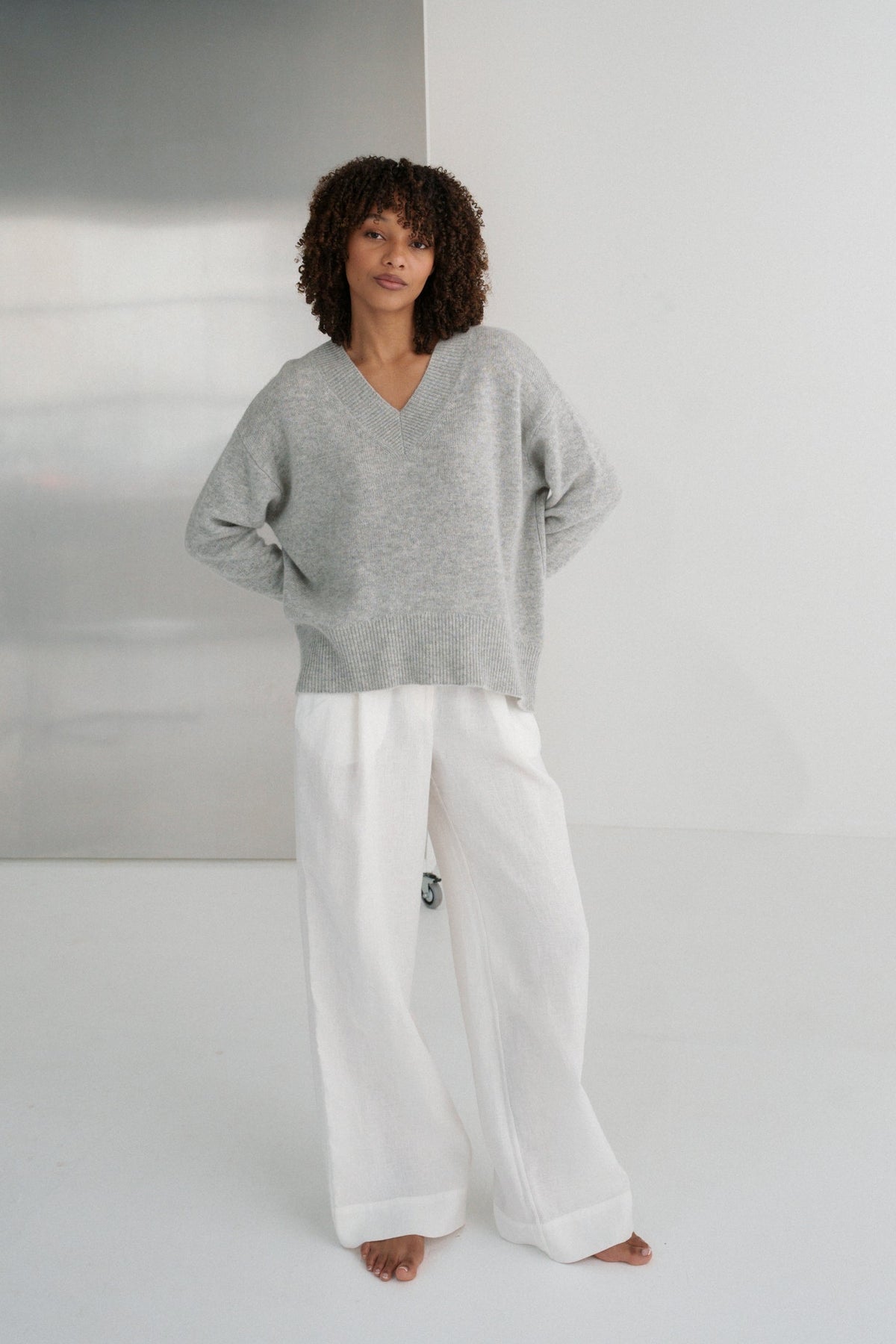 LILLY PILLY woman wearing a grey marle June cashmere knit sweater and ivory linen pants  standing against a white wall with hands on hips