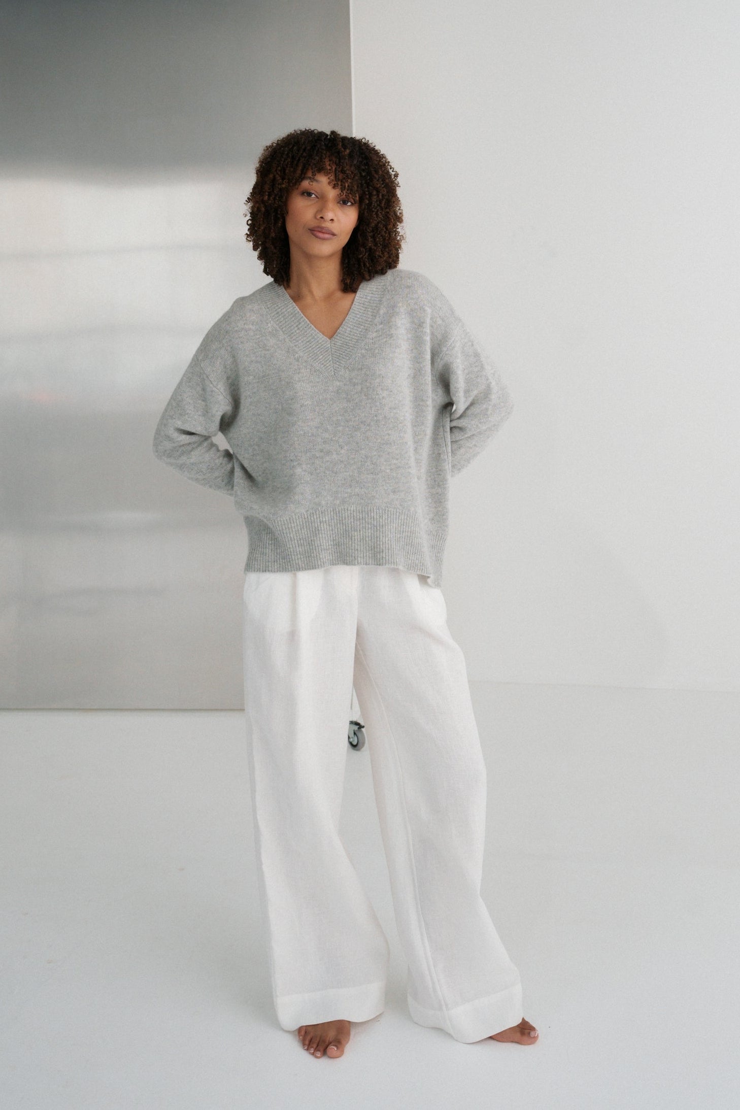 LILLY PILLY woman wearing a grey marle June cashmere knit sweater and ivory linen pants  standing against a white wall with hands on hips
