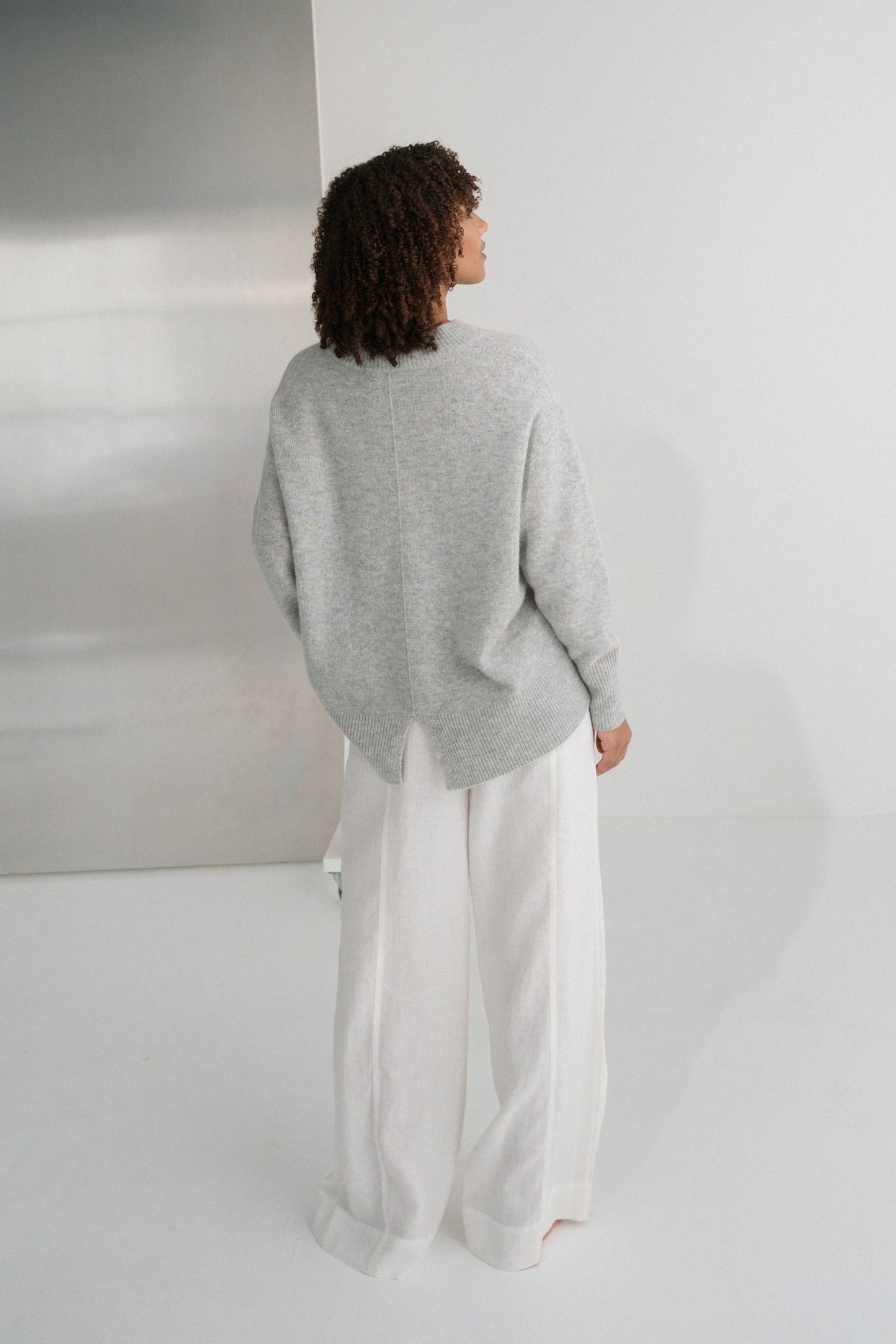 Back view of LILLY PILLY woman wearing a grey marle June cashmere knit sweater and ivory linen pants standing in a minimalistic room with a large mirror.