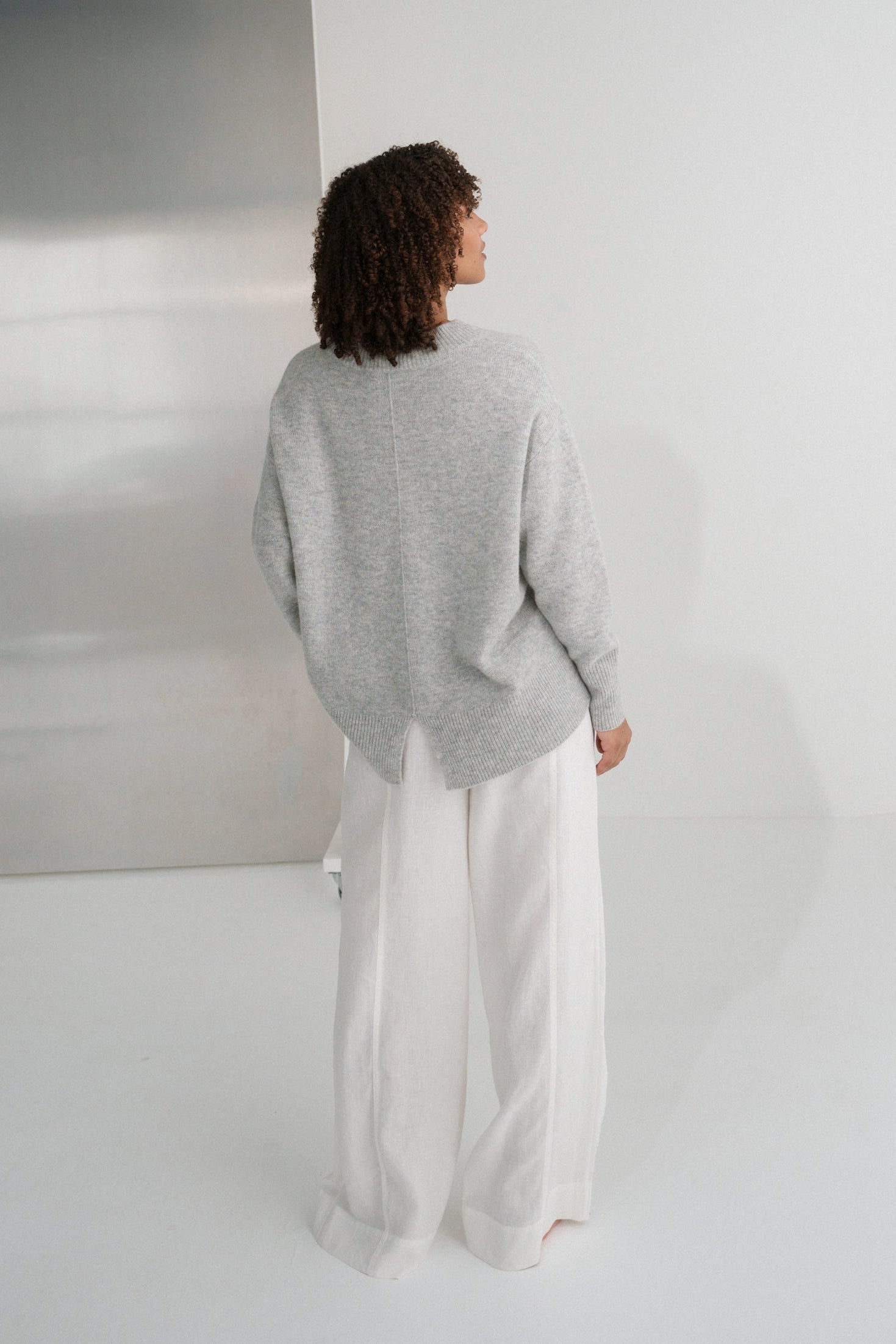 Back view of LILLY PILLY woman wearing a grey marle June cashmere knit sweater and ivory linen pants standing in a minimalistic room with a large mirror.