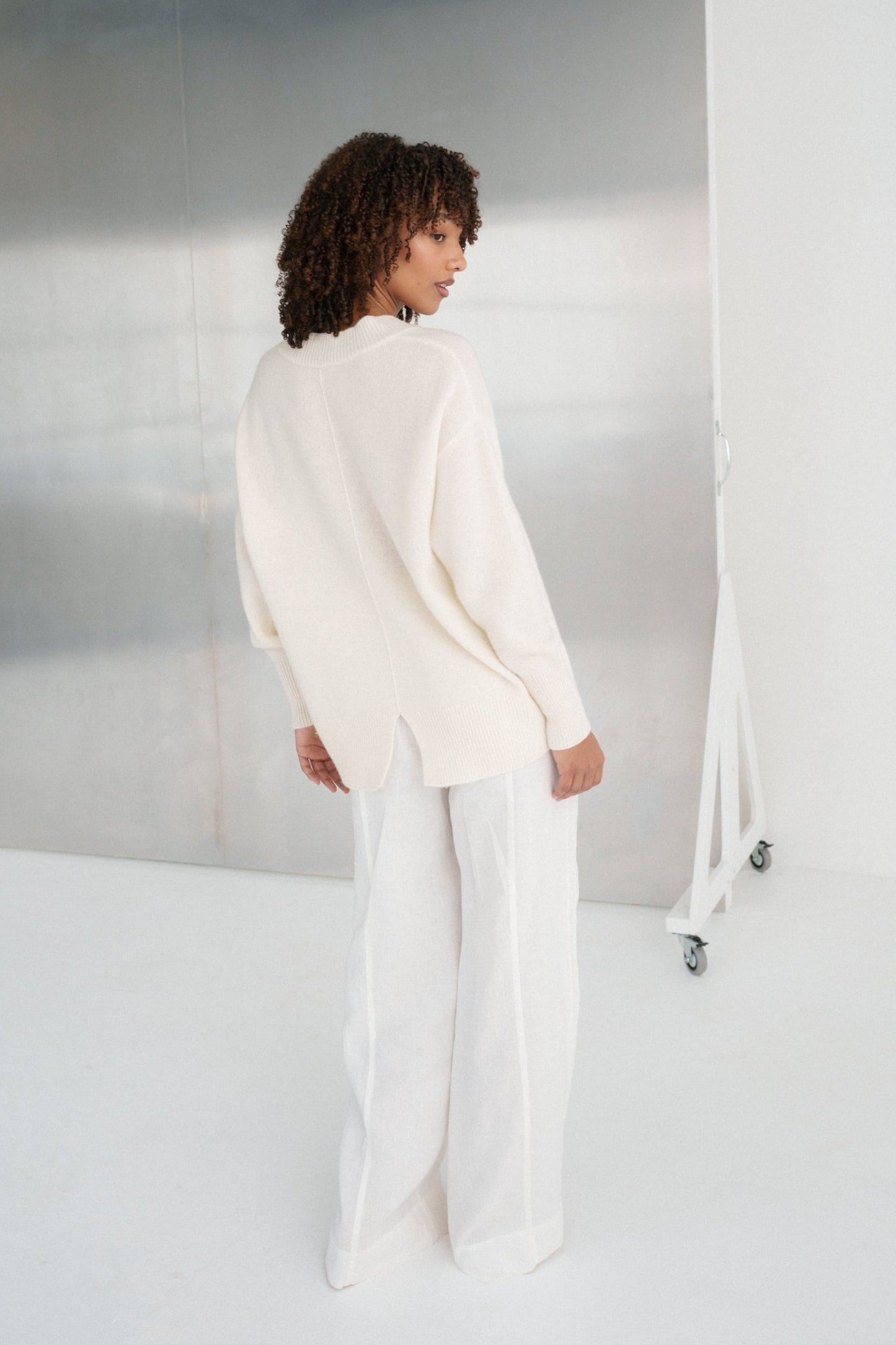 LILLY PILLY woman wearing an ivory June cashmere knit sweater standing in a minimalistic room with a large mirror.