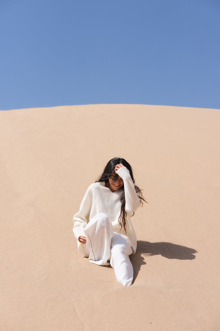 June Cashmere Knit in Ivory - front view, styled with Rika ivory linen pants, seated on a sand dune under a clear blue sky - Lilly Pilly Collection