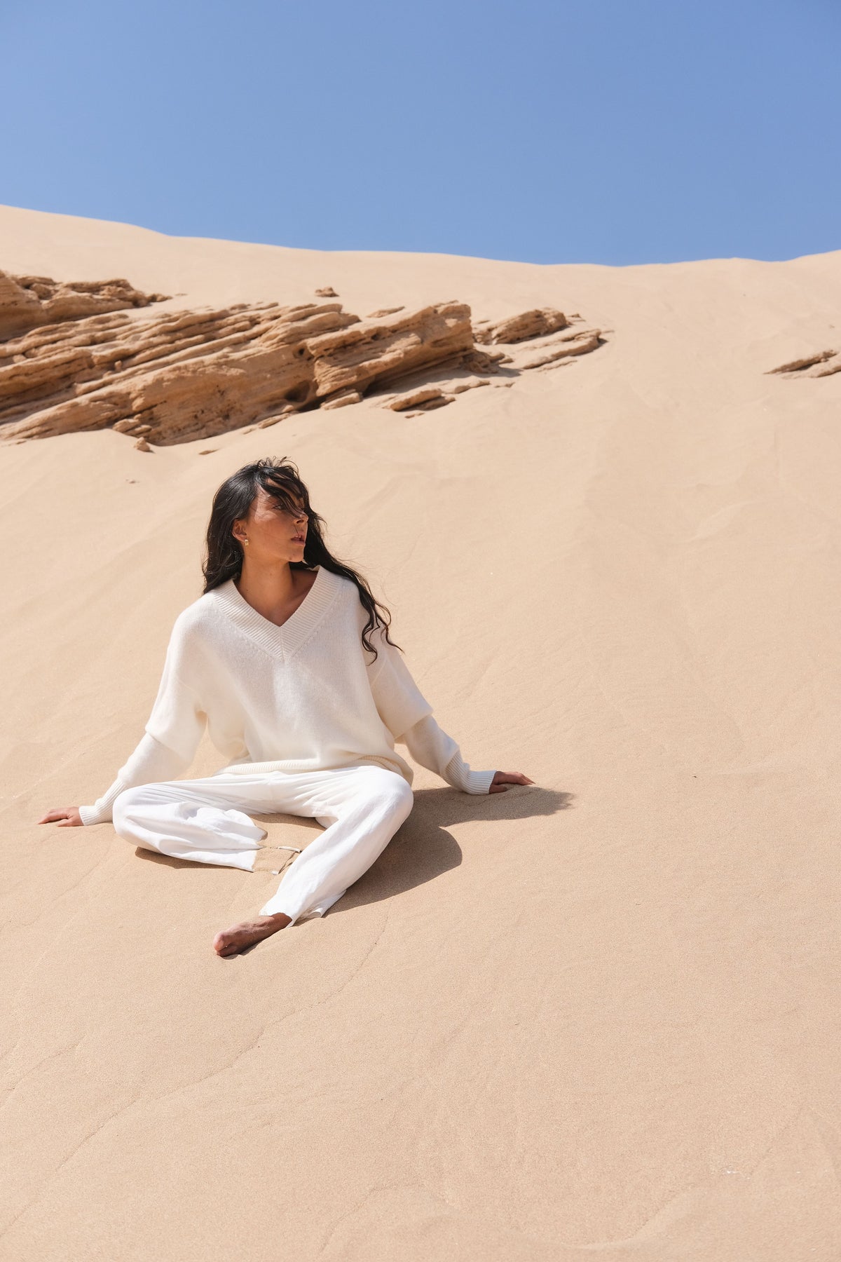 LILLY PILLY woman wearing an ivory June cashmere knit sweater and Rika ivory linen pants outfit sitting on a sand dune with clear blue sky