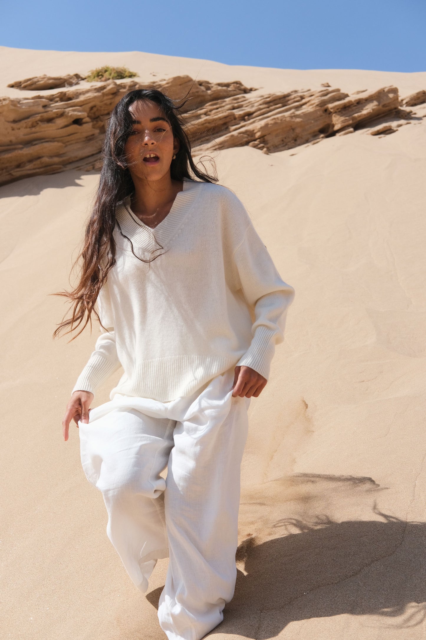 LILLY PILLY woman wearing an ivory June cashmere knit sweater and Rika ivory linen pants outfit running  down a sand dune with a clear blue sky