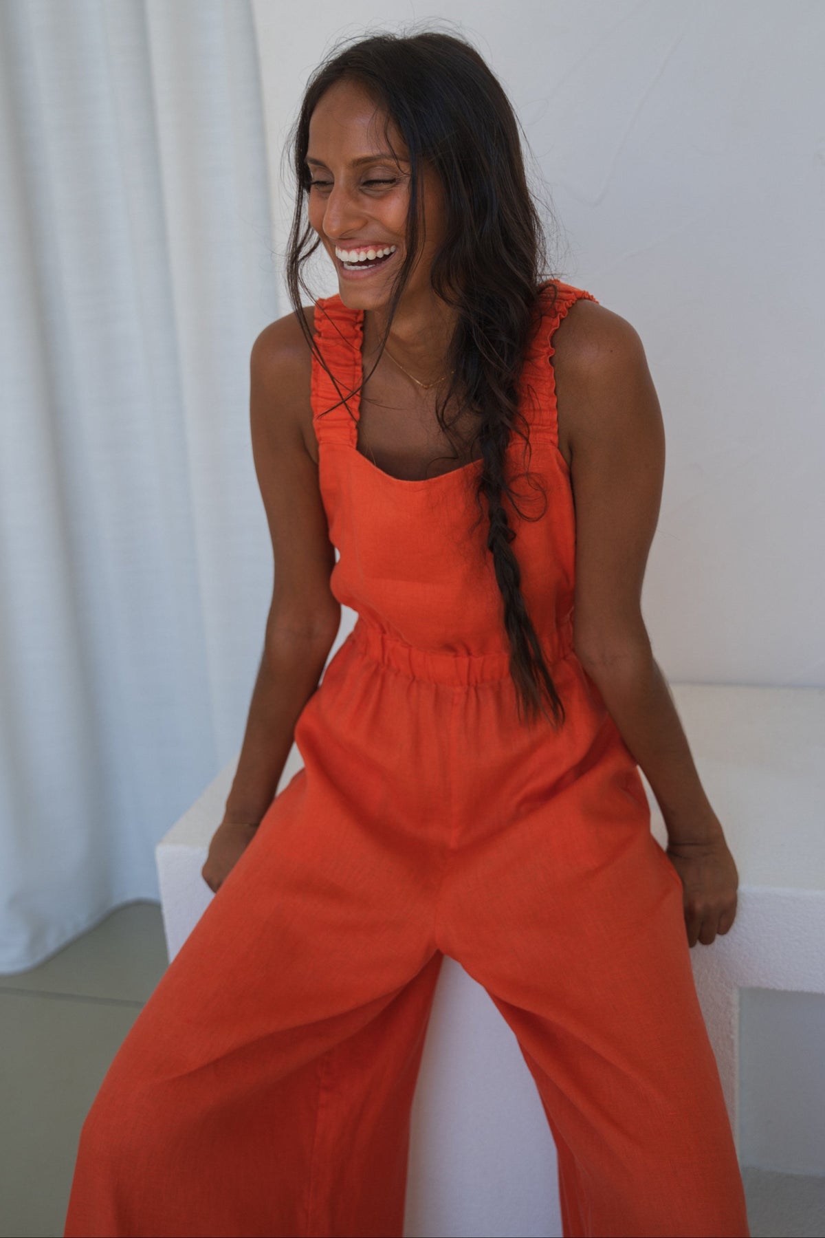 LILLY PILLY woman in an spritz Keely linen jumpsuit sitting on a bench seat with a white curtain background