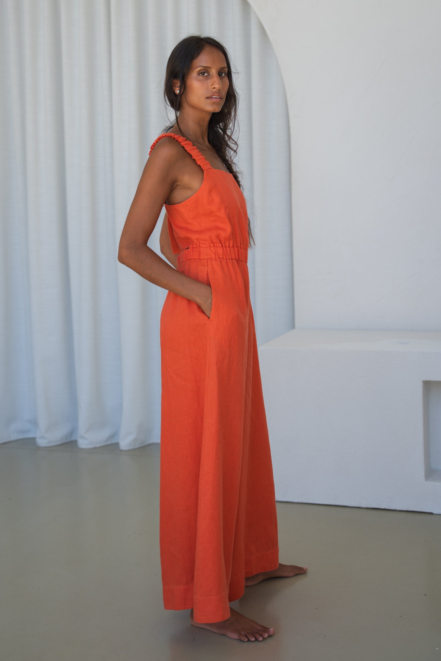 LILLY PILLY woman in an spritz Keely linen jumpsuit side view standing against a white curtain background