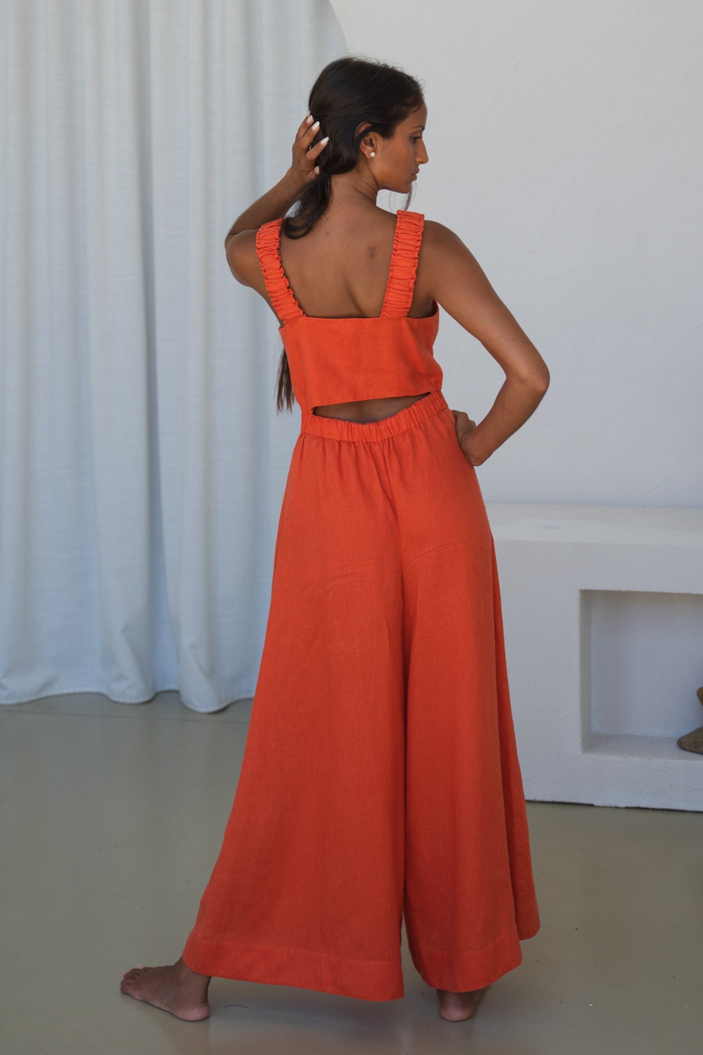 LILLY PILLY woman in an spritz Keely linen jumpsuit showing her back in front of a white curtain background