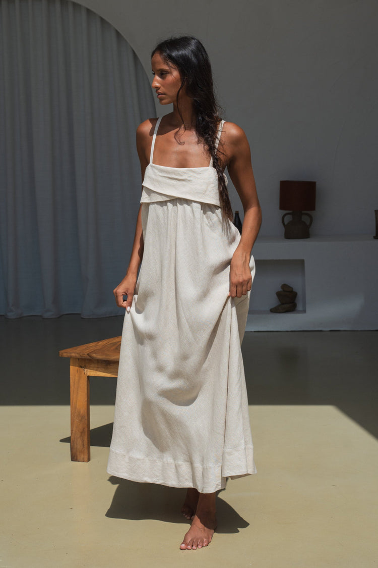 Kiko Linen Midi Dress in Oatmeal - front view, holding skirt fabric, draped neckline, standing near wooden bench - Lilly Pilly Collection