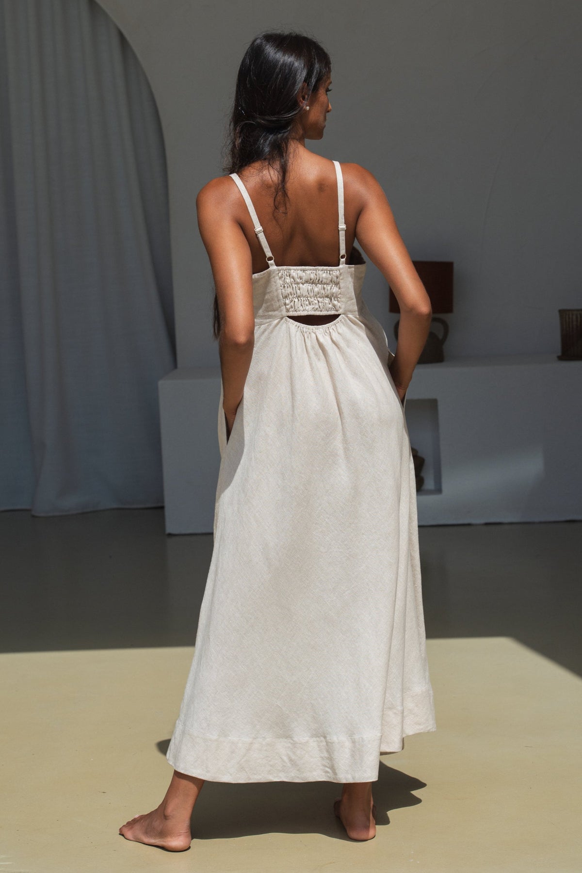 lilly pilly woman in an oatmeal Kiko linen dress standing in a room with thin straps in a minimal indoor setting