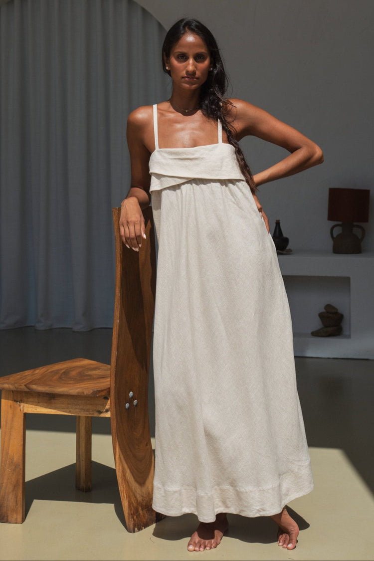 Kiko Linen Midi Dress in Oatmeal - front view, draped asymmetric neckline and spaghetti straps, leaning against wooden chair - Lilly Pilly Collection