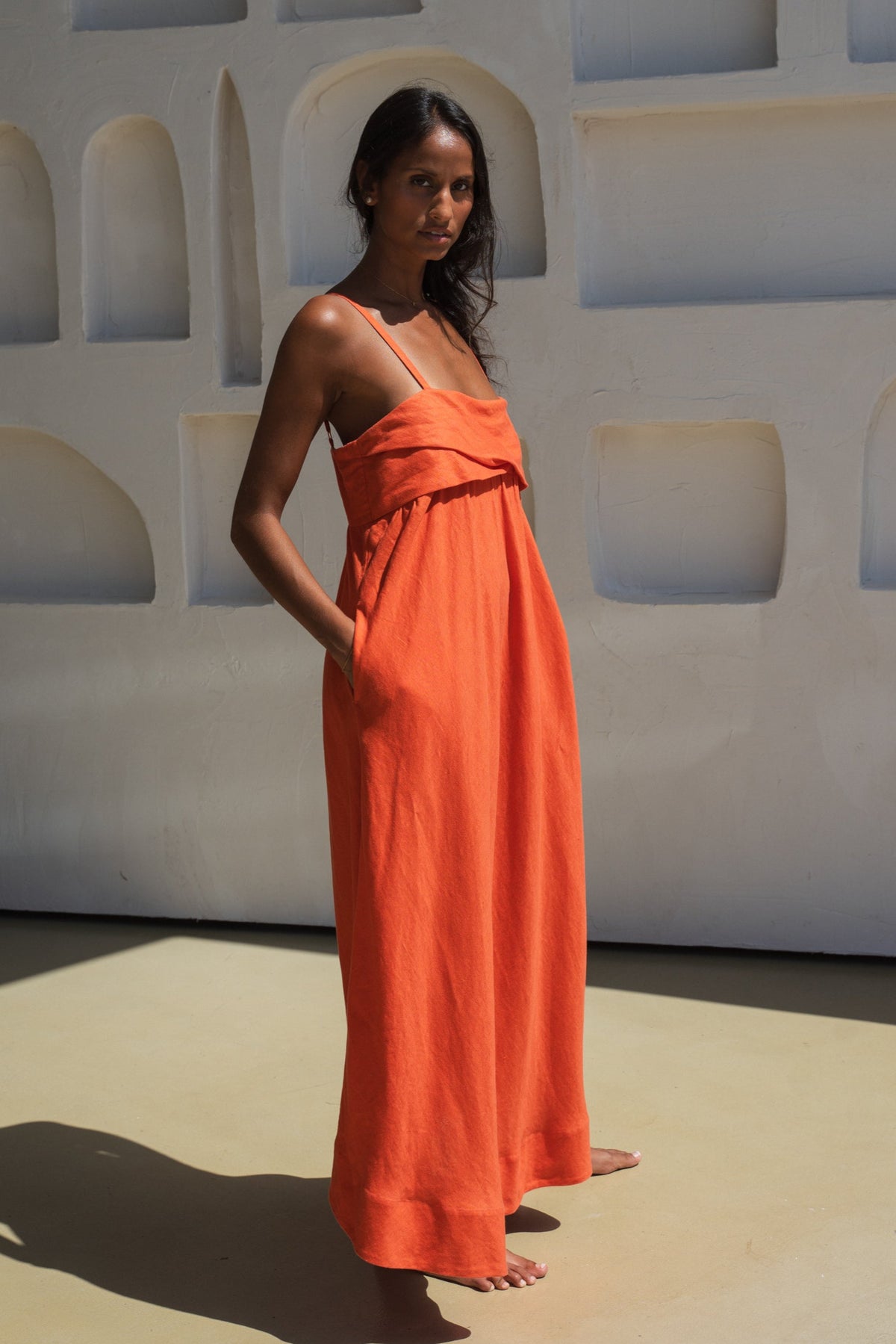 lilly pilly woman showing side view of a spritz coloured Kiko linen dress standing against a white wall with geometric shapes.