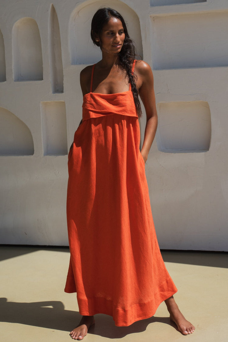 Kiko Linen Midi Dress in Spritz Orange - front view, draped asymmetric neckline and spaghetti straps, standing against textured wall - Lilly Pilly Collection
