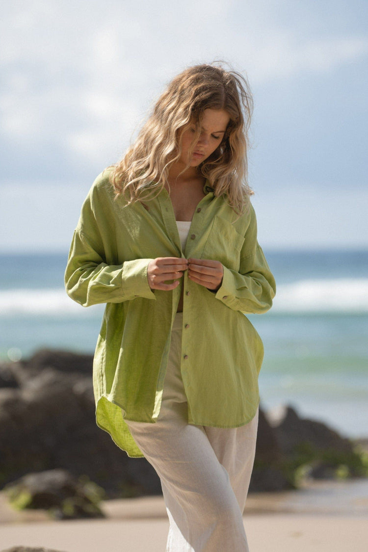 Kirra Linen Shirt in Lemongrass - front view, buttoning, worn open over swimwear, worn with wide-leg trousers, walking on beach with ocean backdrop - Lilly Pilly Collection