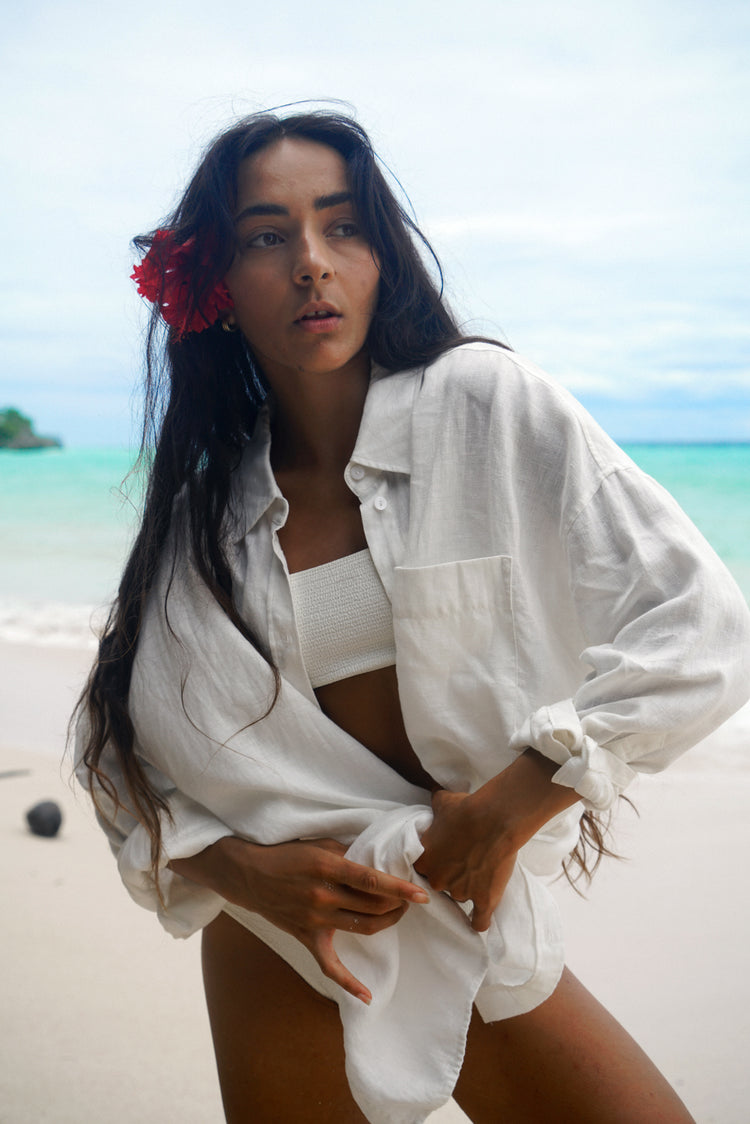 Kirra Linen Shirt in Ivory - worn open and tied at the front over a bikini, tropical beach backdrop with a red flower in the hair - Lilly Pilly Collection