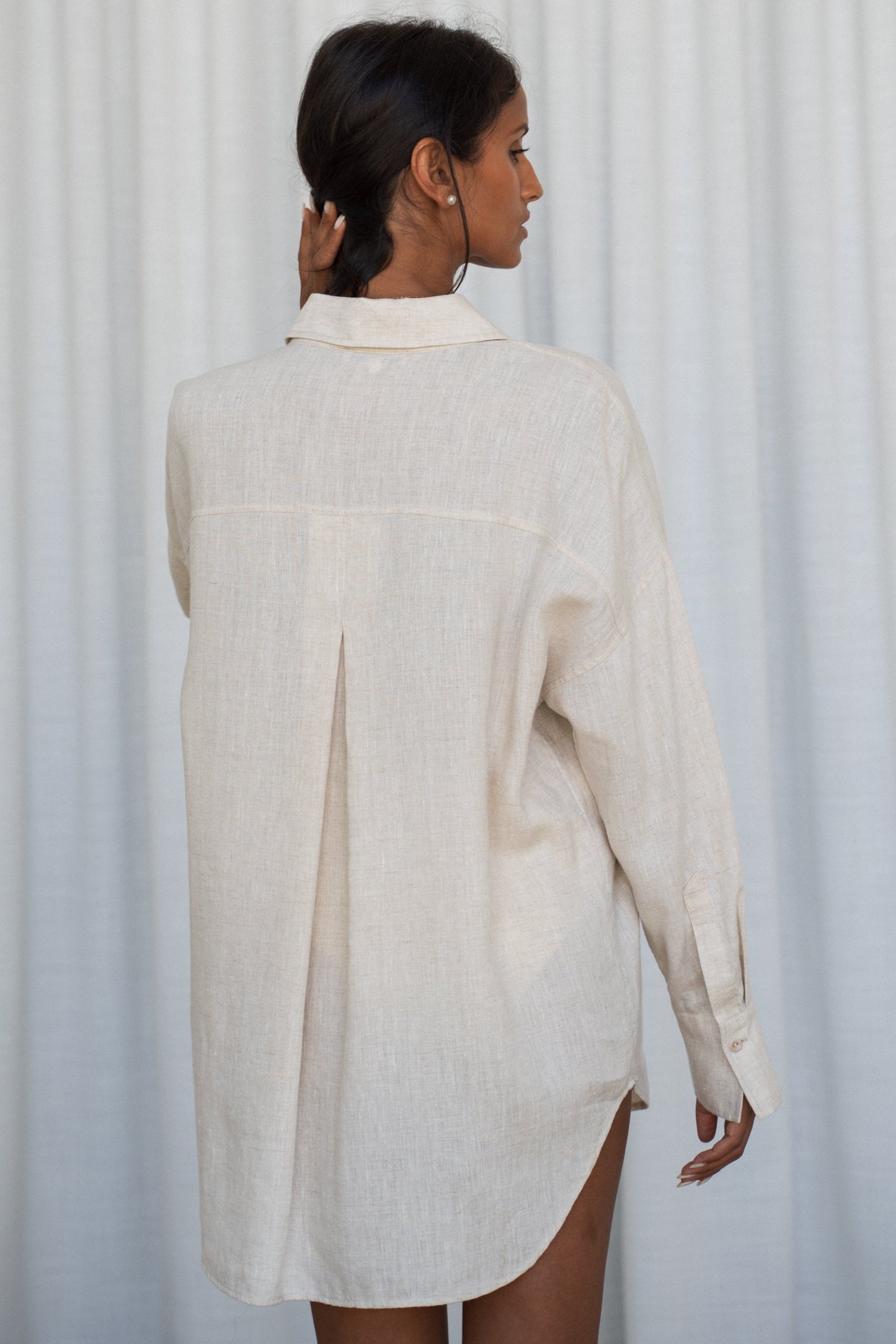 Woman wearing a oatmeal Kirra linen shirt, back view, against a white curtain background