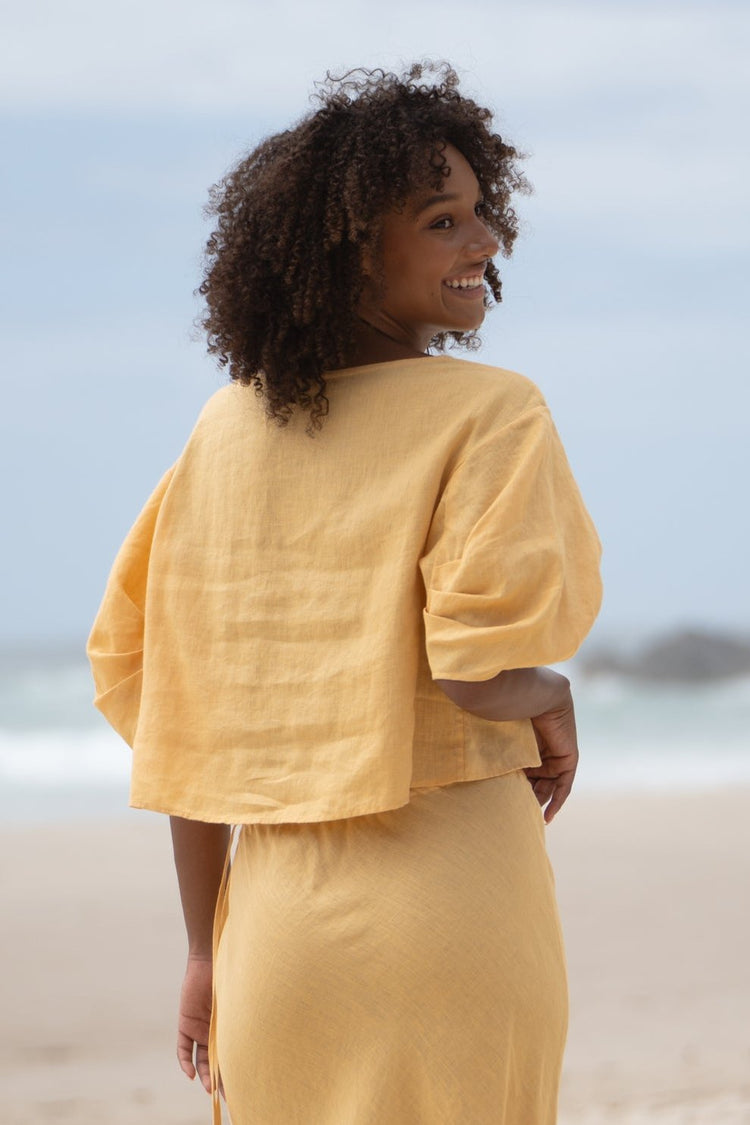Leia Linen Top in Butter Yellow - back view, smiling over shoulder, puff sleeves, worn with matching skirt, standing on beach - Lilly Pilly Collection