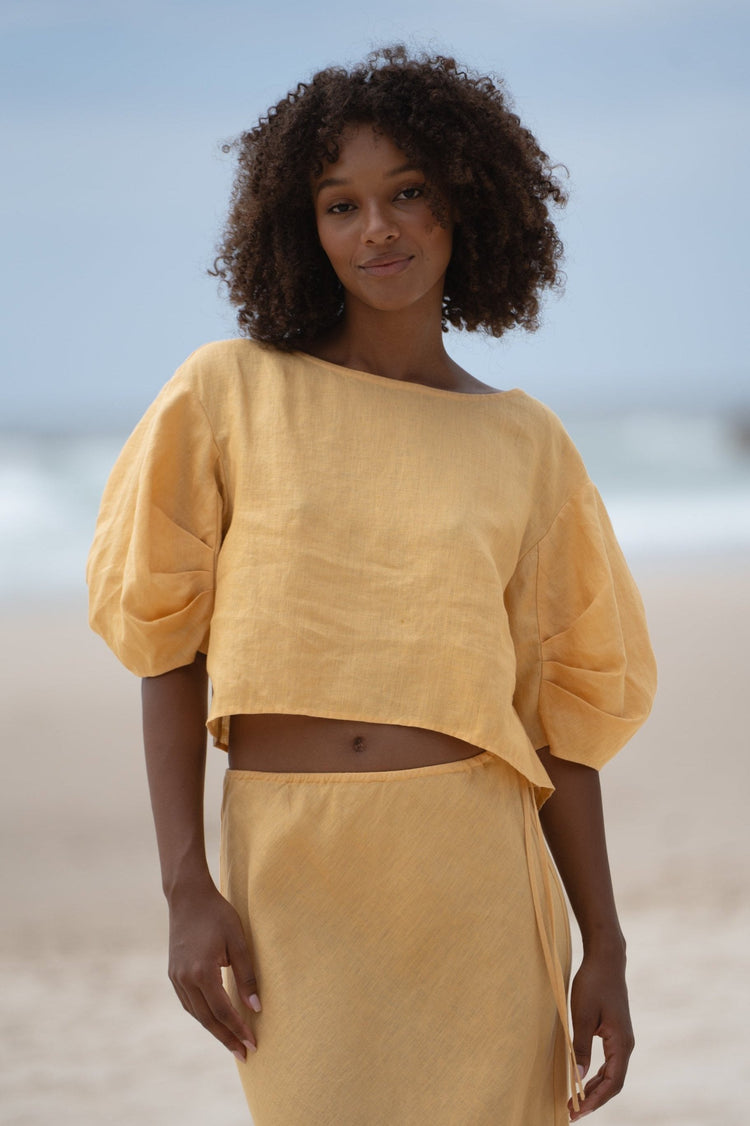 Leia Linen Top in Butter Yellow - front view, off-shoulder boat neckline and puff sleeves, cropped hem, worn with matching skirt, standing on beach - Lilly Pilly Collection