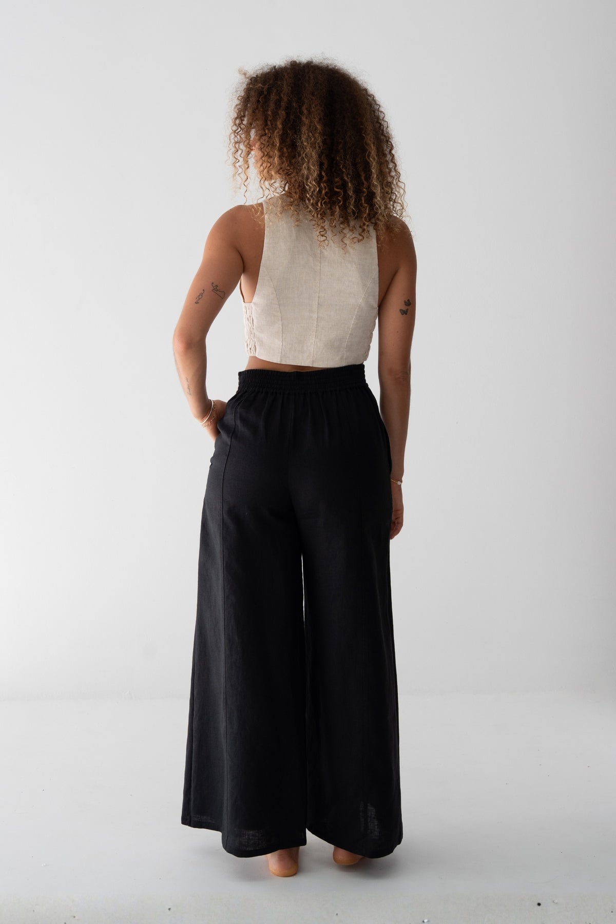 Letty Linen Pants in Black - back view showing elasticated waistband and wide-leg linen silhouette, woman wearing ivory linen waistcoat, studio backdrop - Lilly Pilly Collection