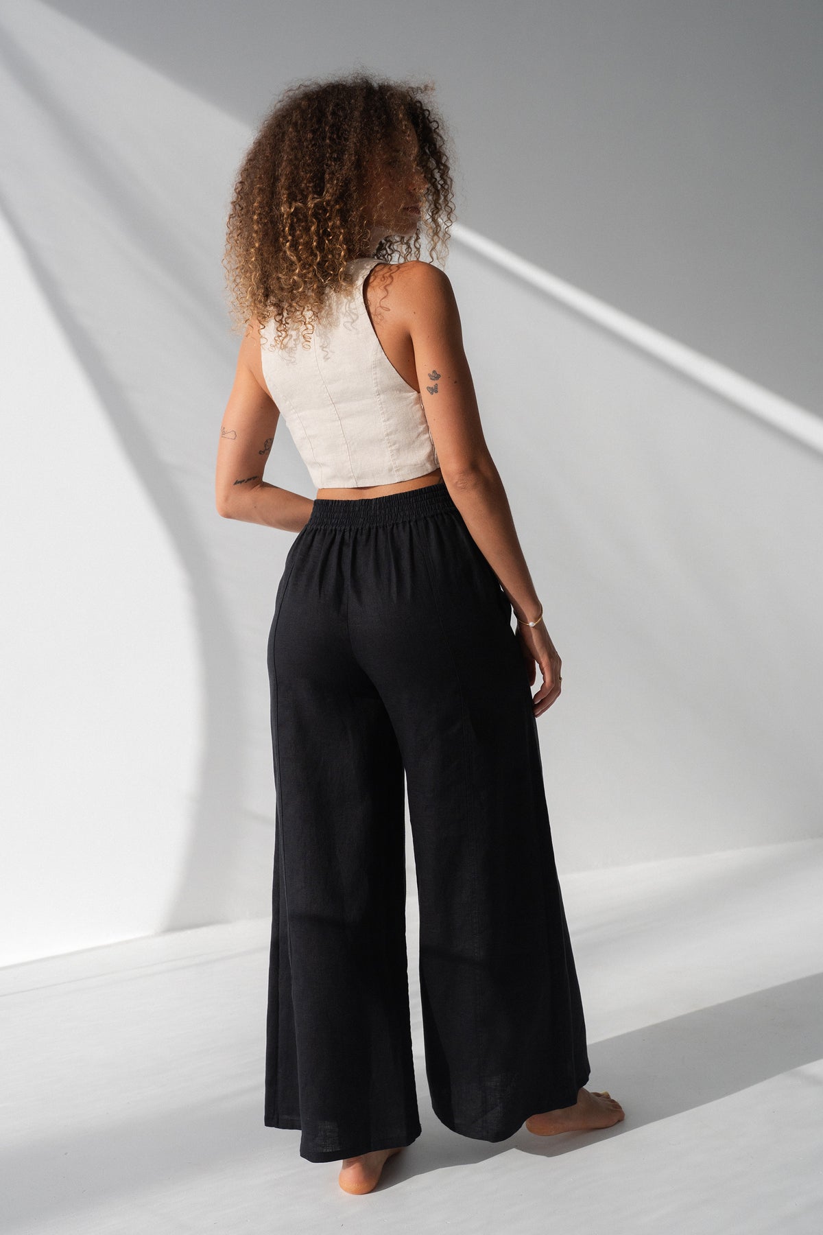 Letty Linen Pants in Black - back view showing elasticated waistband and wide-leg linen drape, woman wearing ivory linen waistcoat, studio backdrop - Lilly Pilly Collection