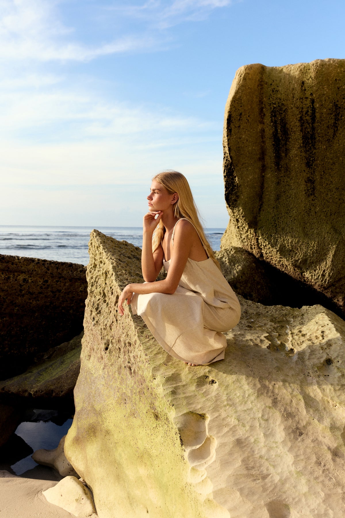 Lia Linen Midi Slip Dress in Oatmeal - woman seated on beach rocks, relaxed midi silhouette, ocean backdrop - Lilly Pilly Collection