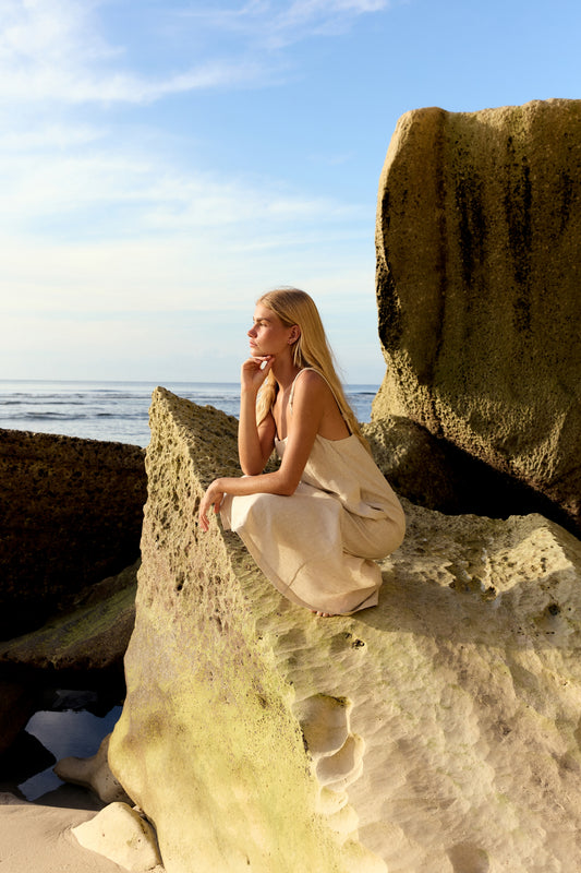 Lia Linen Midi Slip Dress in Oatmeal - woman seated on beach rocks, relaxed midi silhouette, ocean backdrop - Lilly Pilly Collection