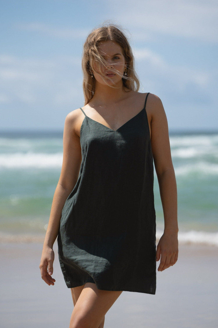 Liz Linen Mini Slip Dress in Bottle Green - front view, walking, V-neckline and thin straps, standing on beach with ocean backdrop - Lilly Pilly Collection