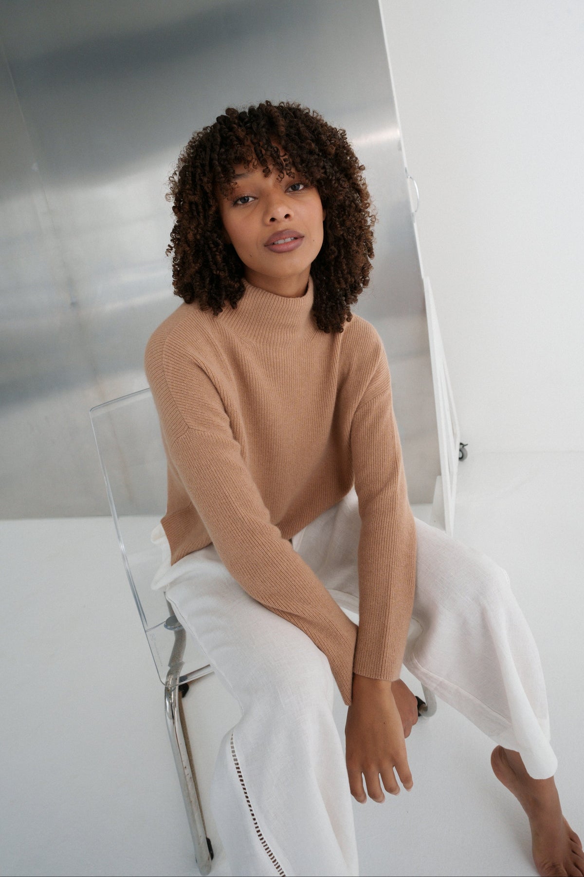 Close up of LILLY PILLY woman wearing a camel brown June cashmere knit sweater and ivory white linen pants sitting in a minimalistic room