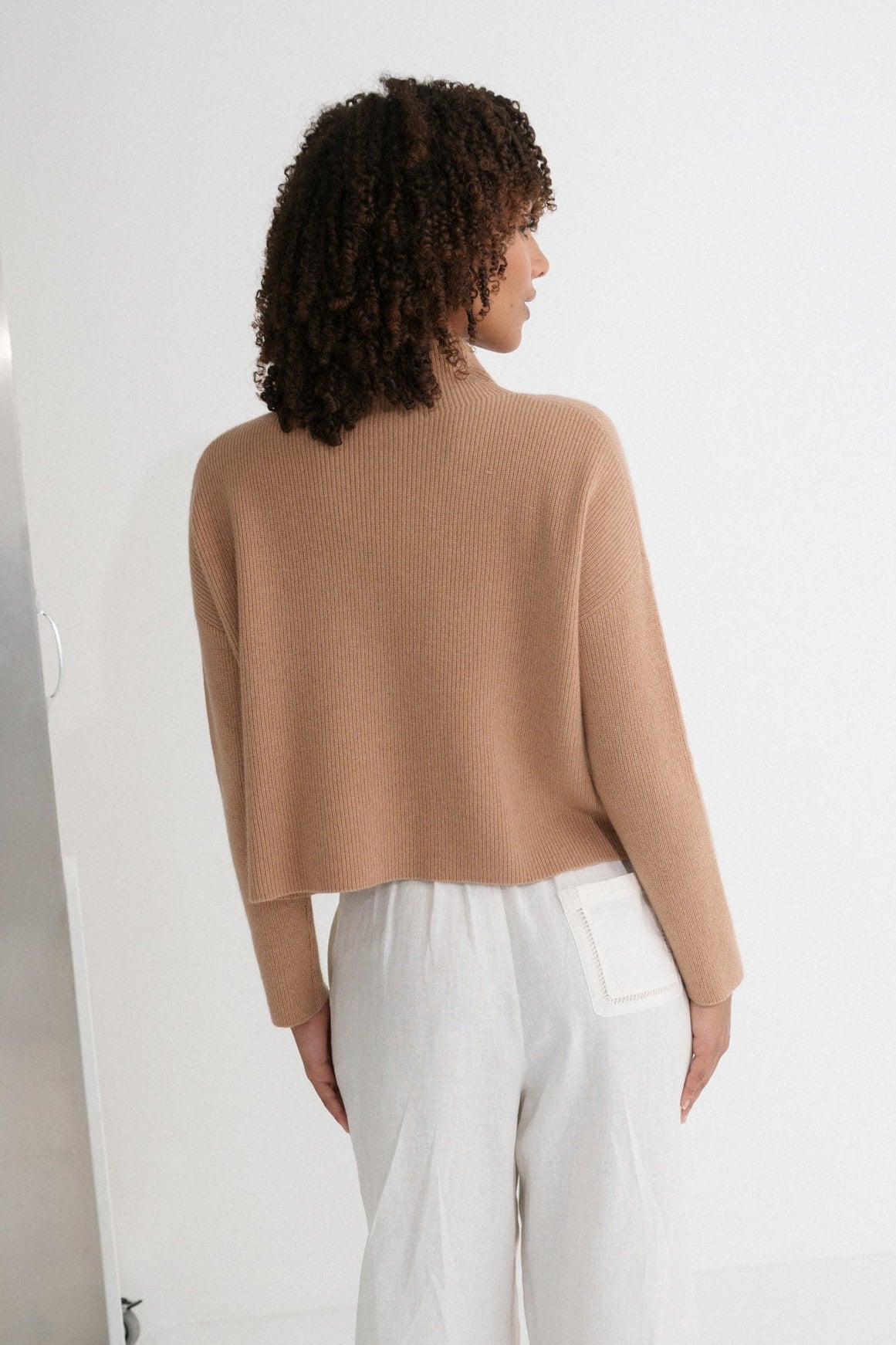 Back view of LILLY PILLY woman wearing a camel brown June cashmere knit sweater and ivory white linen pants standing against a white wall.