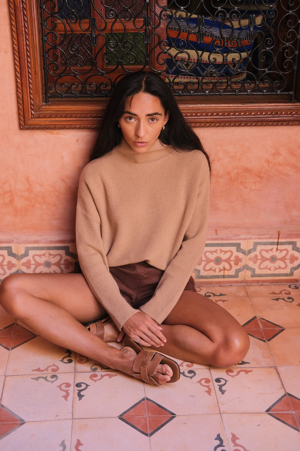 LILLY PILLY woman wearing a camel brown June cashmere knit sweater sitting on a patterned floor wearing a beige sweater and brown shorts.