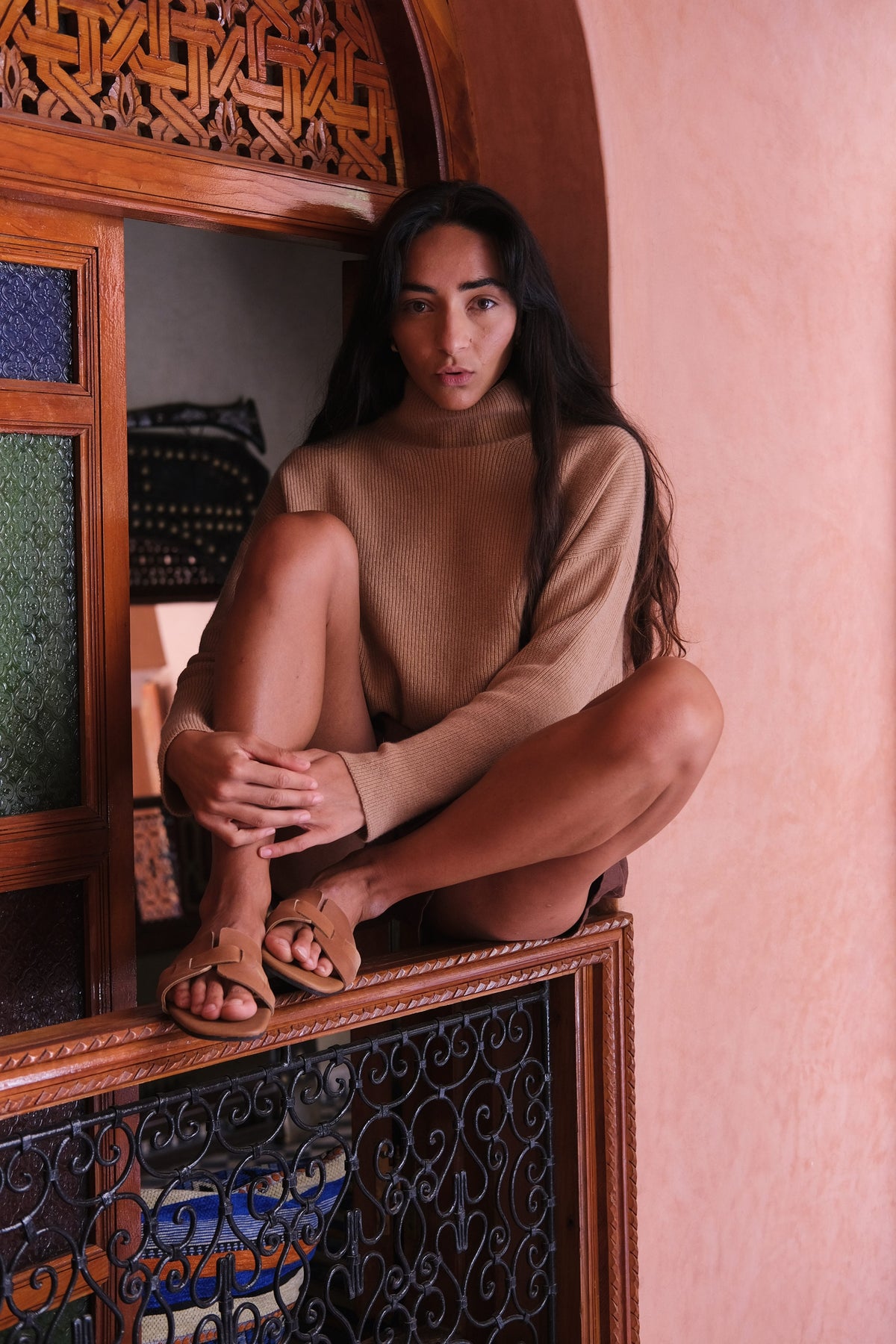 LILLY PILLY woman wearing a camel brown June cashmere knit sweater sitting on a window ledge with ornate wooden and tile work in the background
