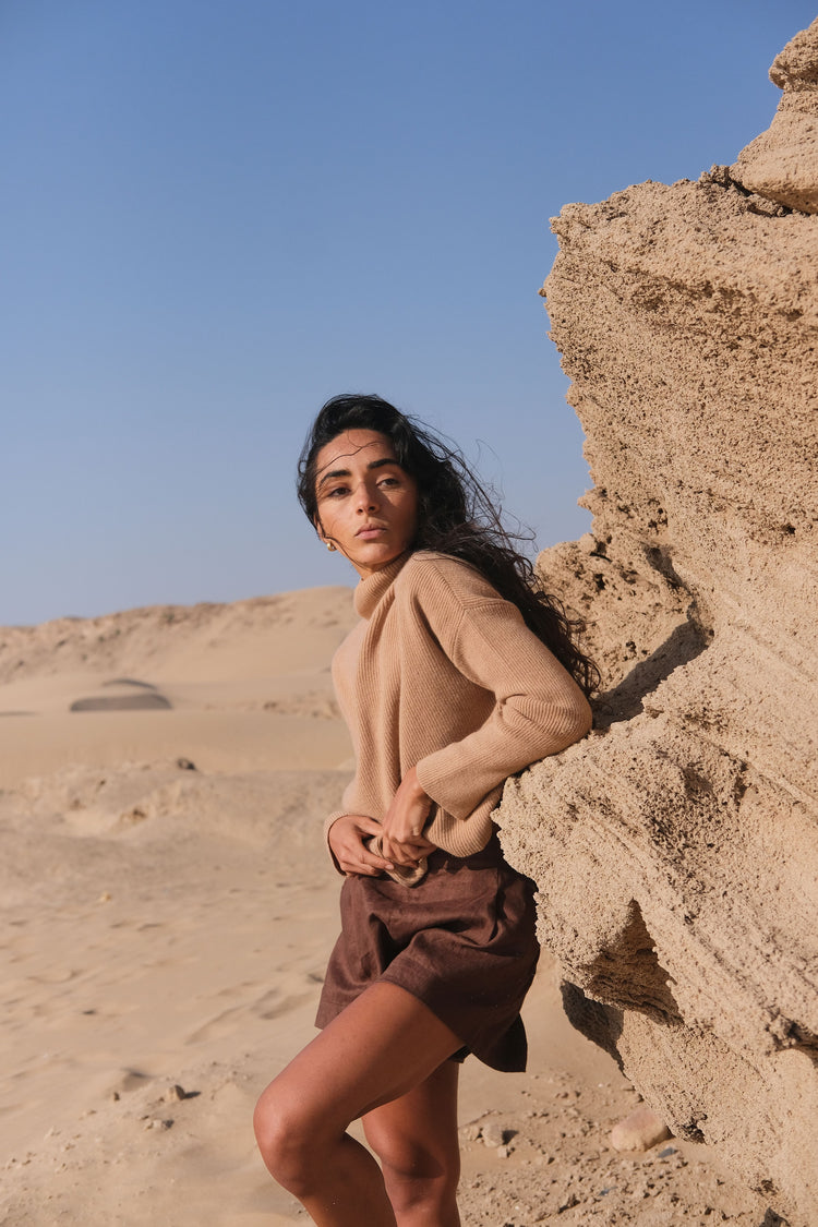 Lumi Cashmere Knit in Camel Brown - front view, standing against a sandstone wall in a desert setting - Lilly Pilly Collection