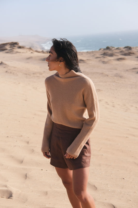 LILLY PILLY woman wearing a camel brown June cashmere knit sweater  standing on a sandy dune with ocean in the background