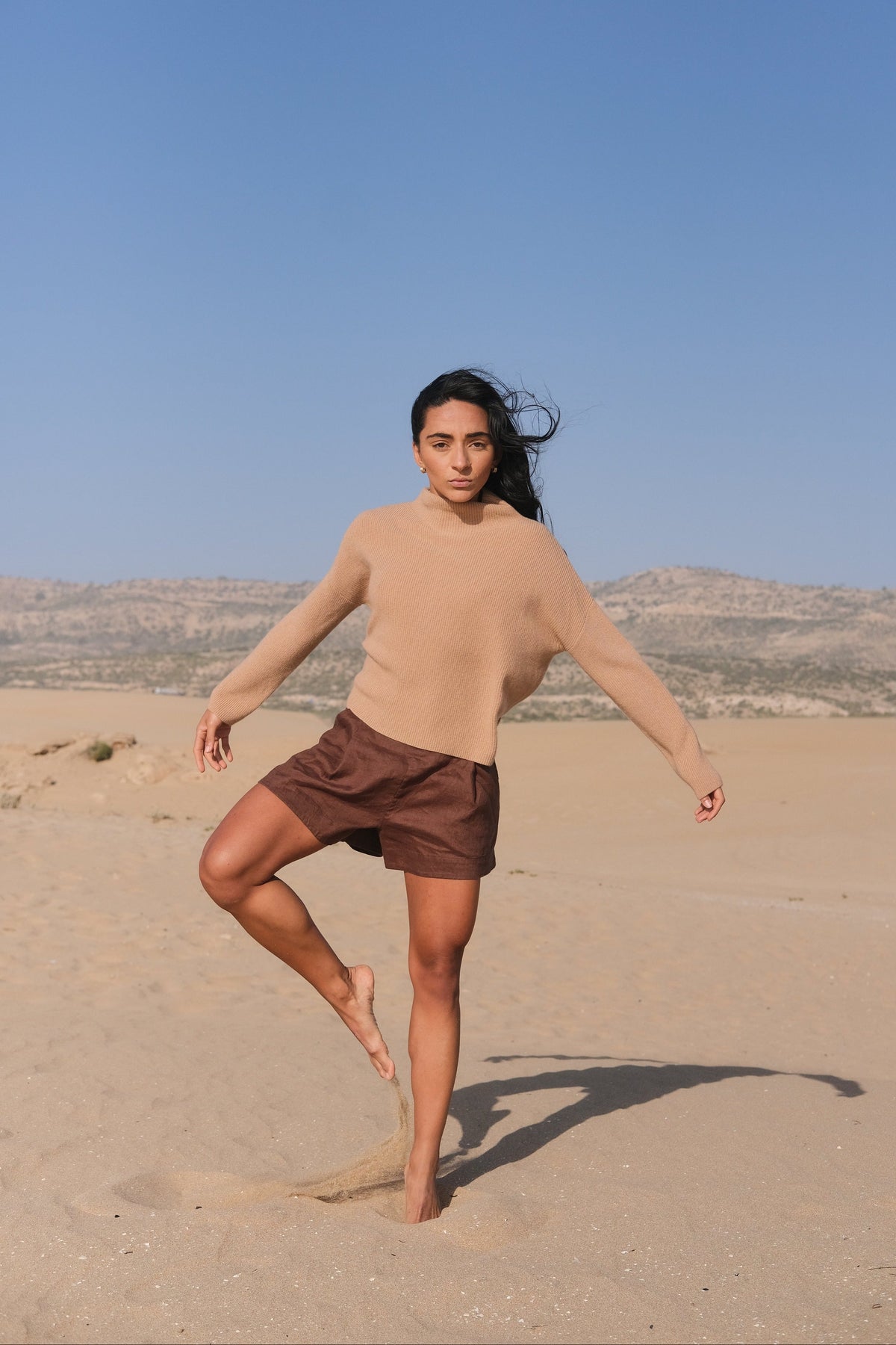 LILLY PILLY woman wearing a camel brown June cashmere knit sweater standing on one leg  in a desert landscape with clear blue sky.
