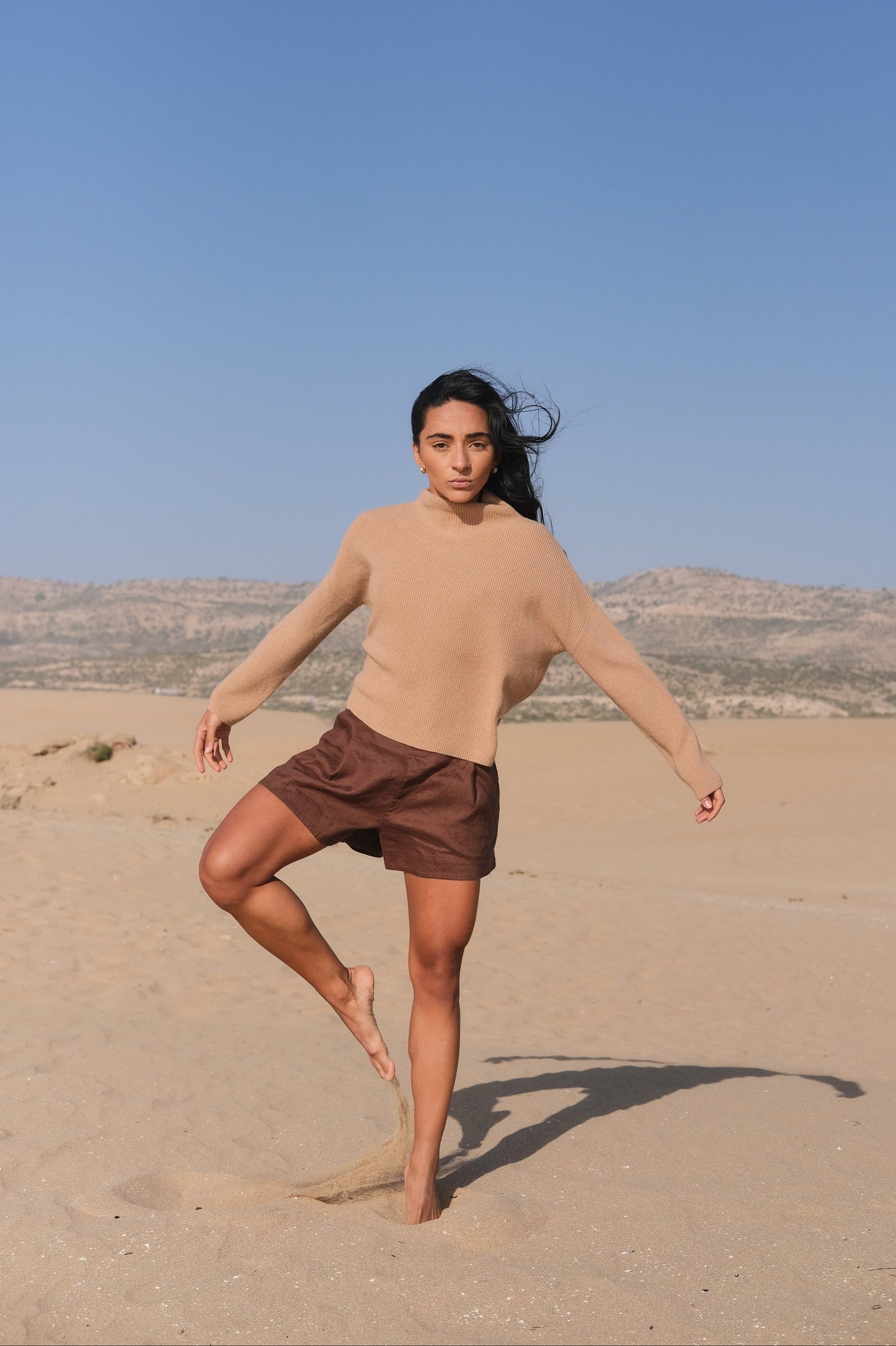 LILLY PILLY woman wearing a camel brown June cashmere knit sweater standing on one leg  in a desert landscape with clear blue sky.